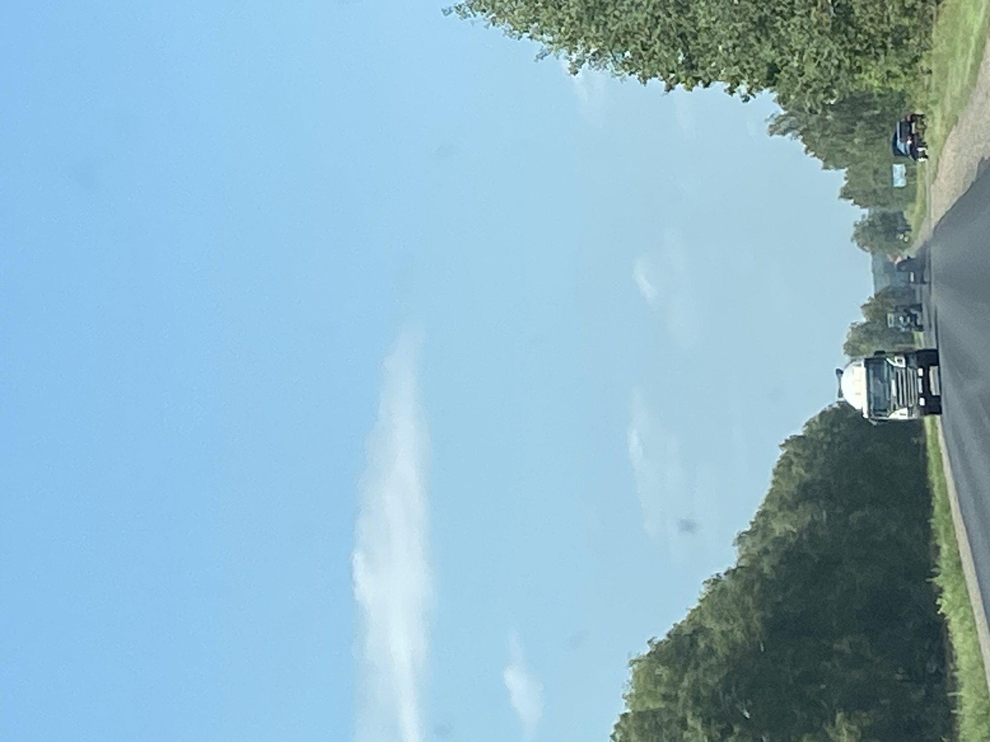 The image depicts a clear, bright day with a vast expanse of blue sky with scattered, wispy clouds. A paved road stretches into the distance, bordered on both sides by lush green trees. Several vehicles are visible on the road. In the foreground, a white truck with a cement mixer is driving towards the viewer. Further down the road, other vehicles, including a tractor and cars, are also visible. A dark-colored car is parked on the side of the road on the right. The overall scene suggests a rural or semi-rural setting in Belarus.
