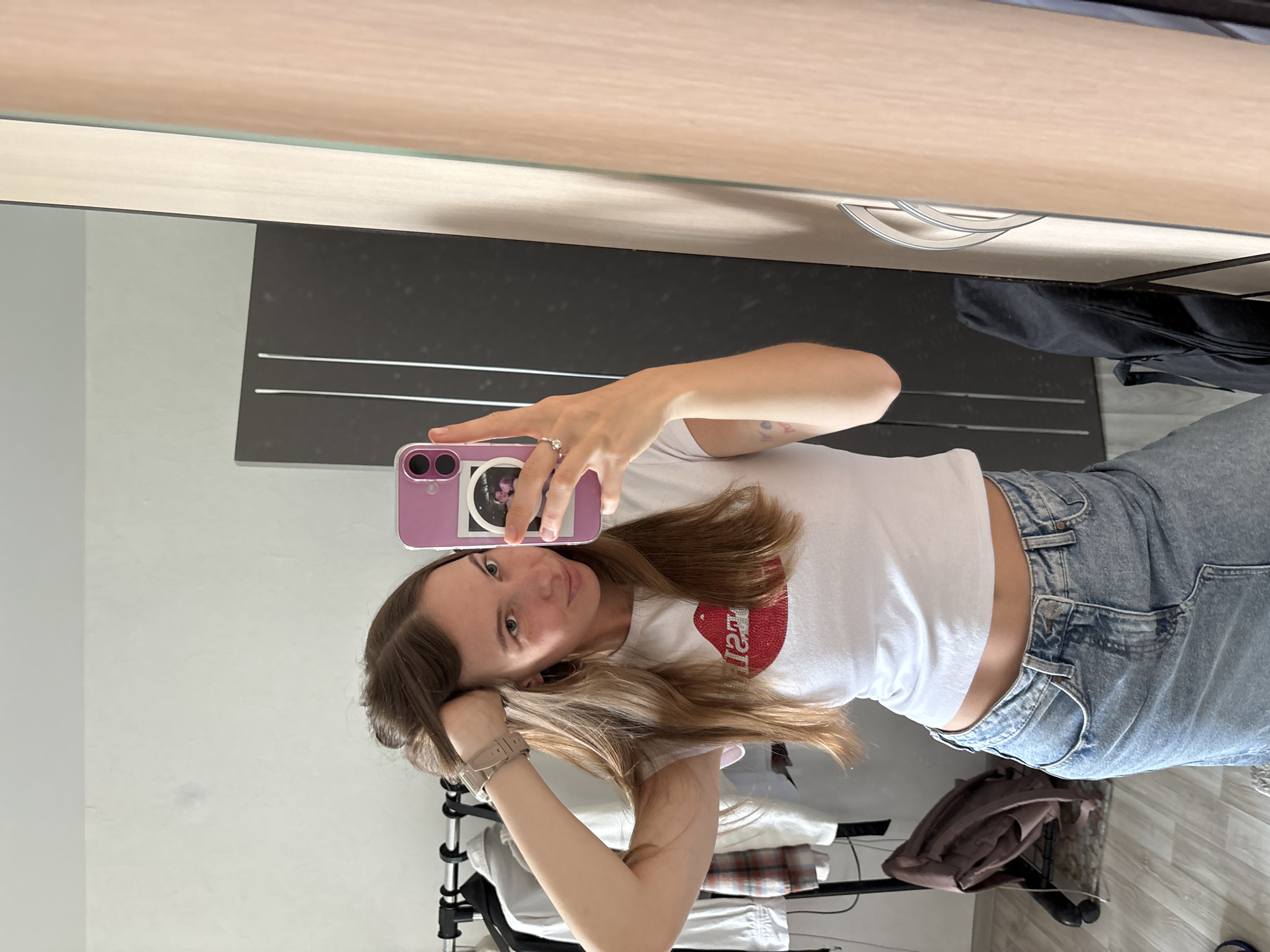 In an indoor setting, possibly a bedroom or dressing area, a young woman with long brown hair stands in front of a full-length mirror, taking a selfie with her phone. She is wearing a white t-shirt with a red lip graphic and a light blue denim skirt. Her right arm is raised, resting her hand in her hair. A watch is visible on her left wrist.
The background reveals a clothing rack with several garments, a dark wardrobe or cabinet, and a portion of what appears to be a light-colored door or paneling. The floor is wooden. The presence of a clothing rack suggests a living space.
No elements in the image explicitly place the scene in Perm, Russia.