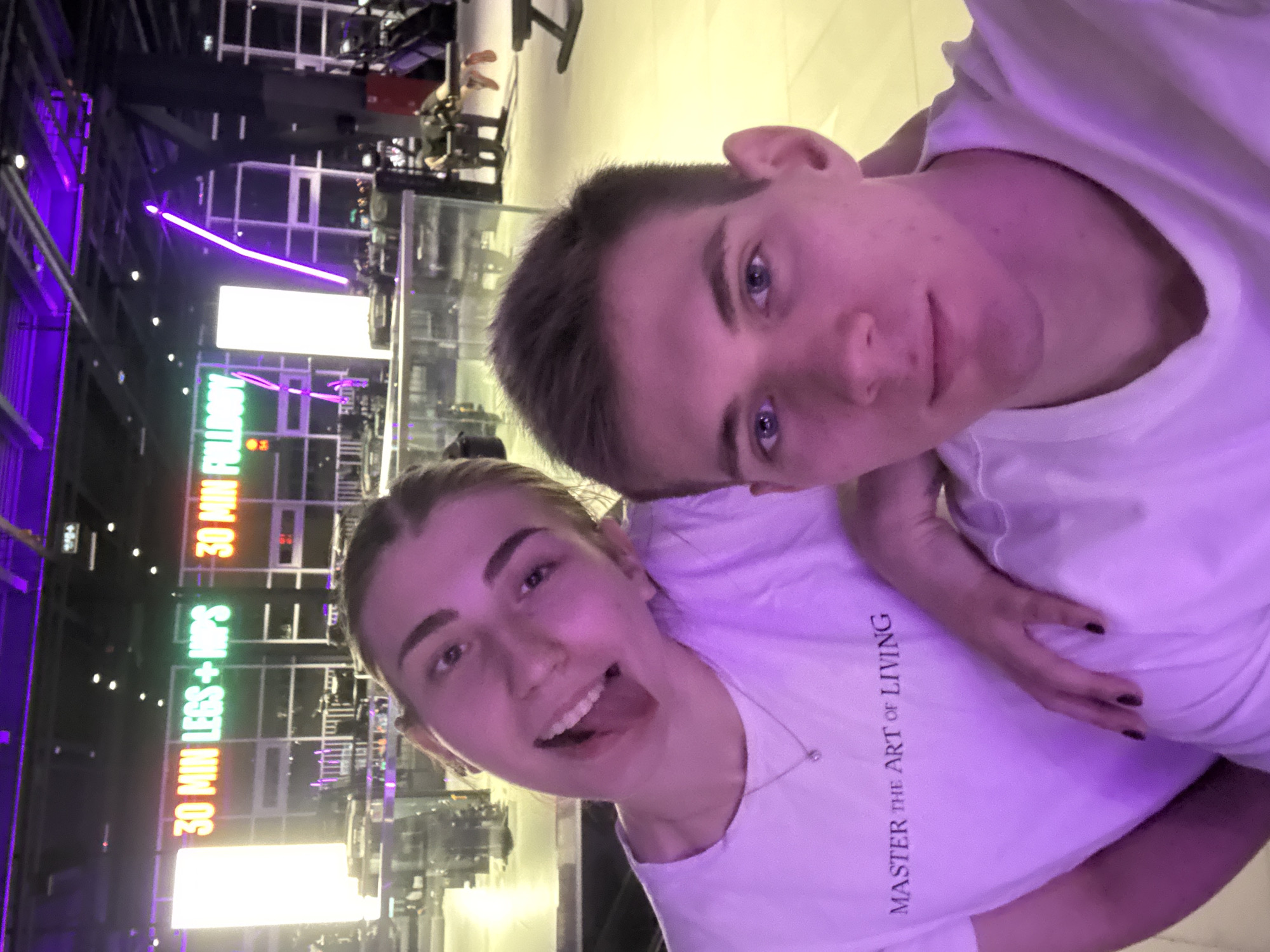 A young couple is taking a selfie in a gym. The woman on the left is sticking her tongue out and smiling. She is wearing a white t-shirt with the text "MASTER THE ART OF LIVING" printed on it. The man on the right is looking directly at the camera with a neutral expression. They are standing in front of a large illuminated sign that says "30 MIN LEGS + HIPS" and "30 MIN FULLBODY". The gym appears to be modern and well-equipped, with various exercise machines visible in the background. The lighting in the gym is a mix of purple and yellow. The location is not specified as Krasnoyarsk, Russia.