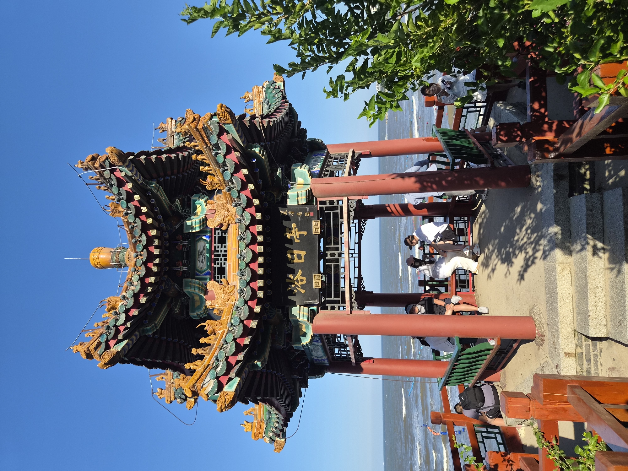An outdoor shot captures a traditional Chinese pavilion by the sea on a clear day. The pavilion features a multi-tiered, ornate roof with green and yellow tiles and intricate carvings, supported by four red pillars. Inscriptions in Chinese characters are visible on the pavilion's facade. Beneath the roof, several people are seated on benches, facing the sea. One person is seated on the left, facing away from the camera, with a backpack. Three people are seated on a bench to the right, facing the sea, two of whom appear to be engaged in conversation. Another person is standing on the far right, looking towards the sea. The sea stretches out to the horizon, with gentle waves and some buoys visible in the water. Lush green foliage is present in the foreground on the right side of the image, casting shadows on the pavilion's floor. The ground leading up to the pavilion is paved with stone steps and a pathway. The overall scene suggests a tourist attraction or a scenic viewpoint. The inscription on the pavilion reads "The Sunbathing Pavilion" (浴日亭). This pavilion is a prominent landmark located on Beidaihe Beach in Qinhuangdao, China, a popular seaside resort destination.