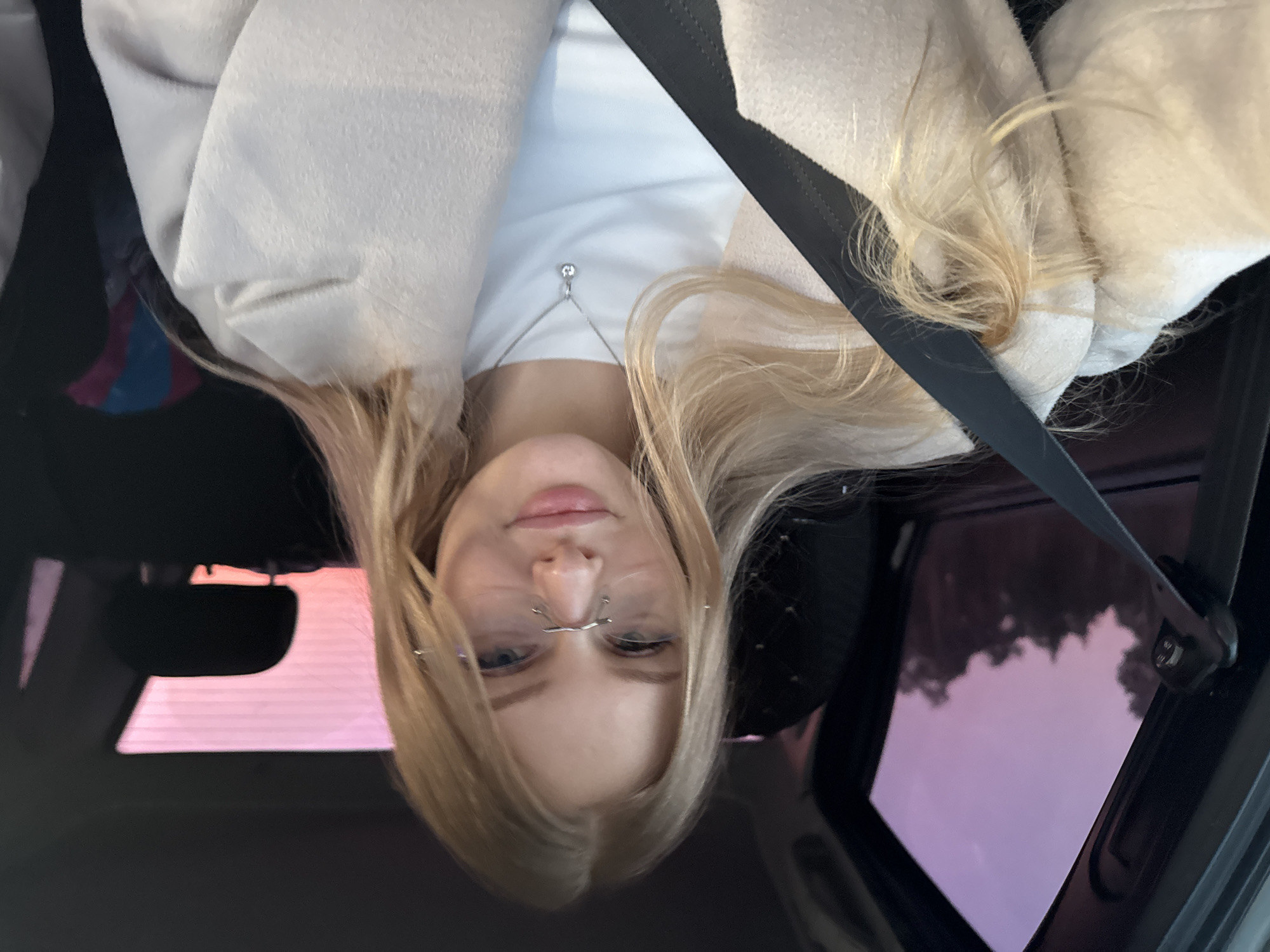 A fair-skinned female with blonde hair sits in a vehicle, wearing a white shirt, a light-colored coat, and a seatbelt. She has a silver nose piercing. The vehicle's open sunroof reveals a sky with pink hues, possibly at sunset or sunrise. The location is potentially in Perm, Russia, but there is no definite evidence.