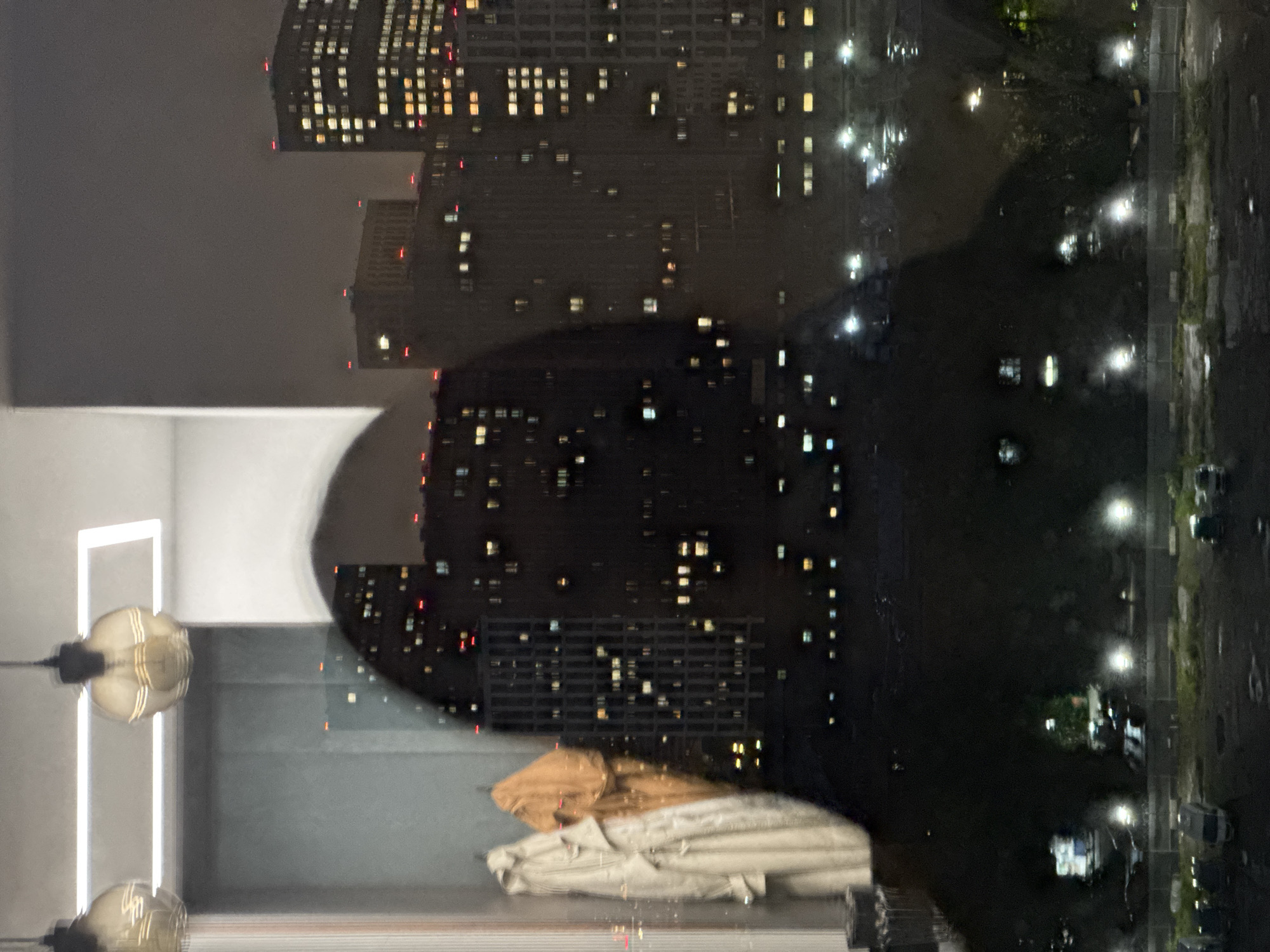 The image depicts a reflection in a window at night. The primary subjects are two light fixtures with visible bulbs, hanging from the ceiling. They are positioned in the foreground and appear to be part of an interior space, possibly a room or hallway, suggested by a white wall and a rectangular light feature behind them.
Reflected in the window is a nighttime cityscape dominated by tall buildings illuminated with numerous lights, indicating a dense urban environment. The buildings have a grid-like pattern of windows, some of which are lit with warm yellow or orange light, while others appear dark. There are also some red lights visible on the tops of some buildings, possibly indicating navigation lights for aircraft. The reflection also shows streetlights casting a glow on what appears to be a street or plaza. The overall impression is one of a busy city at night.
The time of day is clearly night. The weather is not directly discernible, but the reflections and the dimly lit urban environment suggest it might be overcast or that the city lights are reflecting off a slightly humid surface. The location cues, such as the architectural style of the buildings and the dense urban setting, are consistent with a major metropolitan area. Given the context of Moscow, Russia, the scene likely represents a view of the city at night.
There is no discernible text in the image. The overall mood is somewhat muted due to the darkness and the reflections, but the bright city lights offer a sense of life and activity within the urban landscape. The presence of the indoor lighting fixtures contrasted with the expansive cityscape creates an interesting juxtaposition between interior and exterior environments.