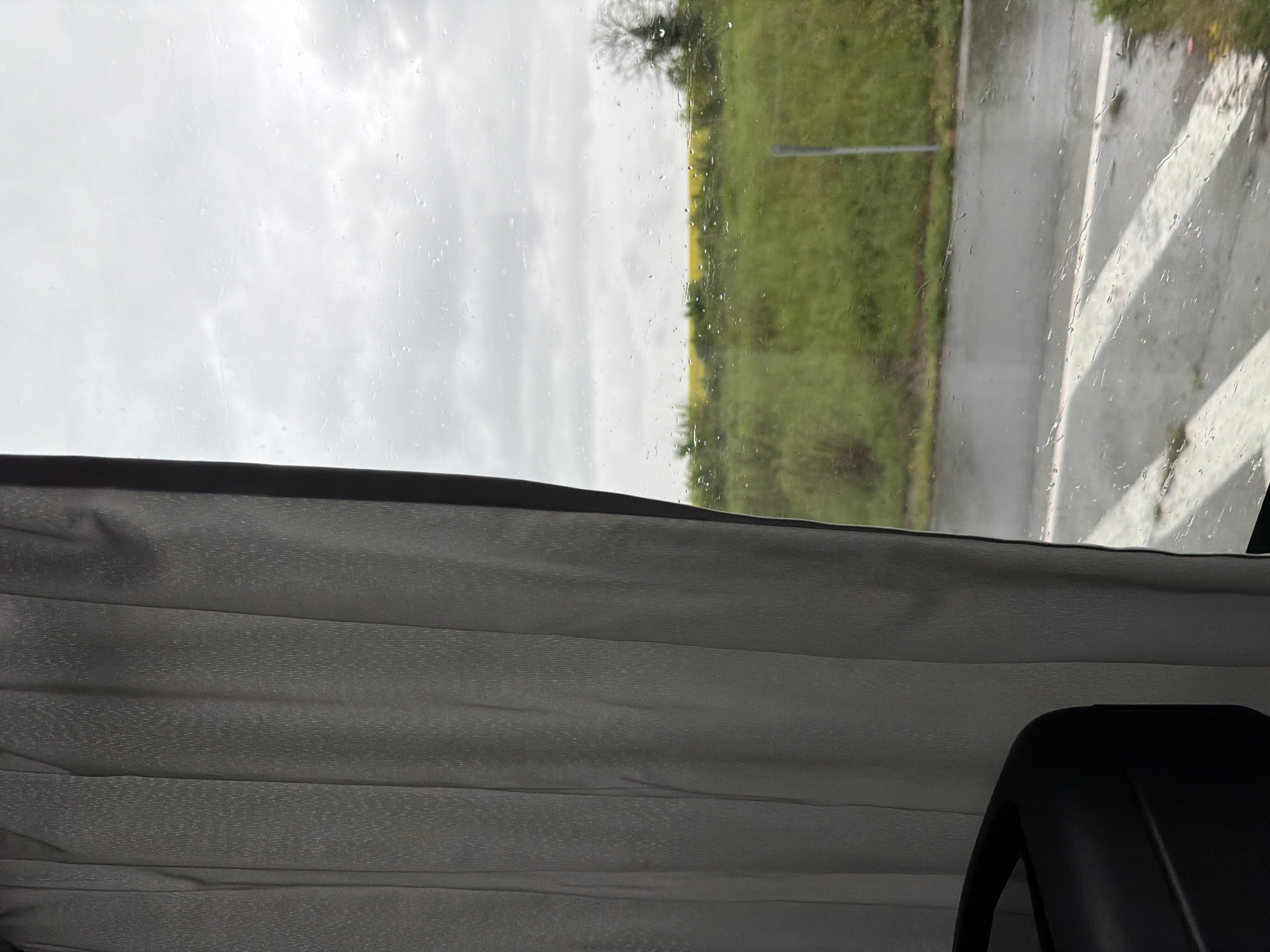The image captures a perspective from inside a vehicle, looking out a side window during an overcast and rainy day. The view is presented as if the camera was held sideways, showing the scene rotated 90 degrees clockwise.
In the lower half of the frame, a pleated grey fabric, resembling a curtain or blind, is visible, indicating the vehicle's interior. A dark, possibly black, object, potentially part of a car seat or interior fixture, is partially visible in the very bottom right.
Through the window, numerous raindrops cling to the glass, suggesting ongoing or recent rainfall. Beyond the wet pane, the sky is uniformly grey and cloudy. The landscape consists of expansive green fields or grasslands, with subtle patches of yellow, possibly from blooming crops like rapeseed. Scattered trees, some appearing bare or with minimal foliage, punctuate the horizon. To the right of the view through the window, a wet, grey road or paved area is visible, marked with distinct white lines, which could be lane markings or a pedestrian crossing. The scene evokes a sense of travel or observation during rainy weather in a rural or semi-rural setting. The location context provided is Rogozinovo, Bulgaria. No text is visible in the image.