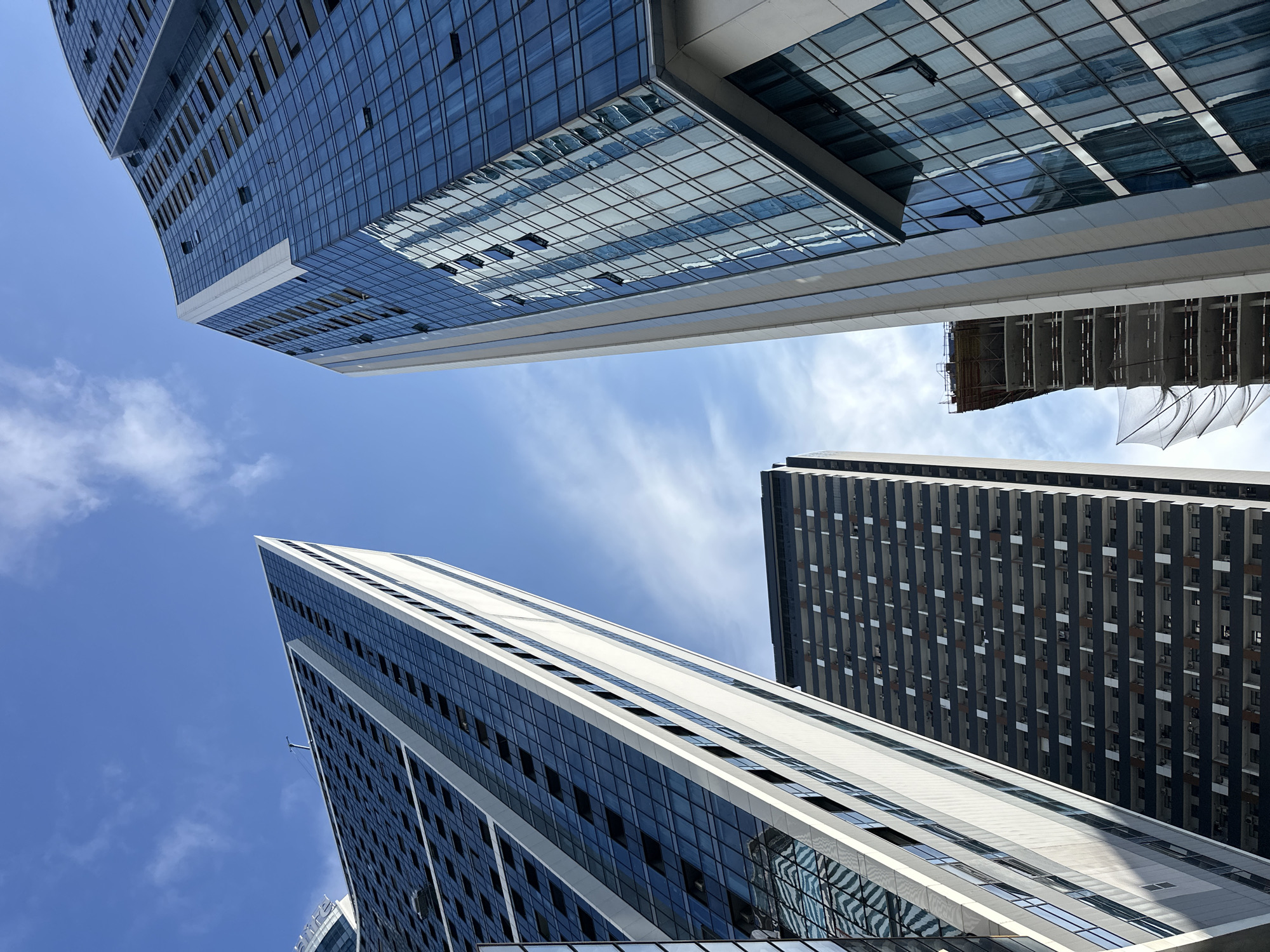 The image shows a low-angle view of several modern high-rise buildings in Batumi, Georgia. The buildings are tall and appear to be residential or commercial. The architecture is characterized by extensive use of glass facades, reflecting the blue sky and clouds. One building on the right has a curved, blue glass exterior, with many windows. To its left, a narrower building with white vertical accents and dark glass panels is angled upwards. In the foreground and center, a darker building with a grid-like pattern of balconies and windows is visible. Further in the background, a building under construction is partially visible, with scaffolding and netting. The sky is clear blue with scattered white clouds. The overall impression is one of urban development and modern architecture.