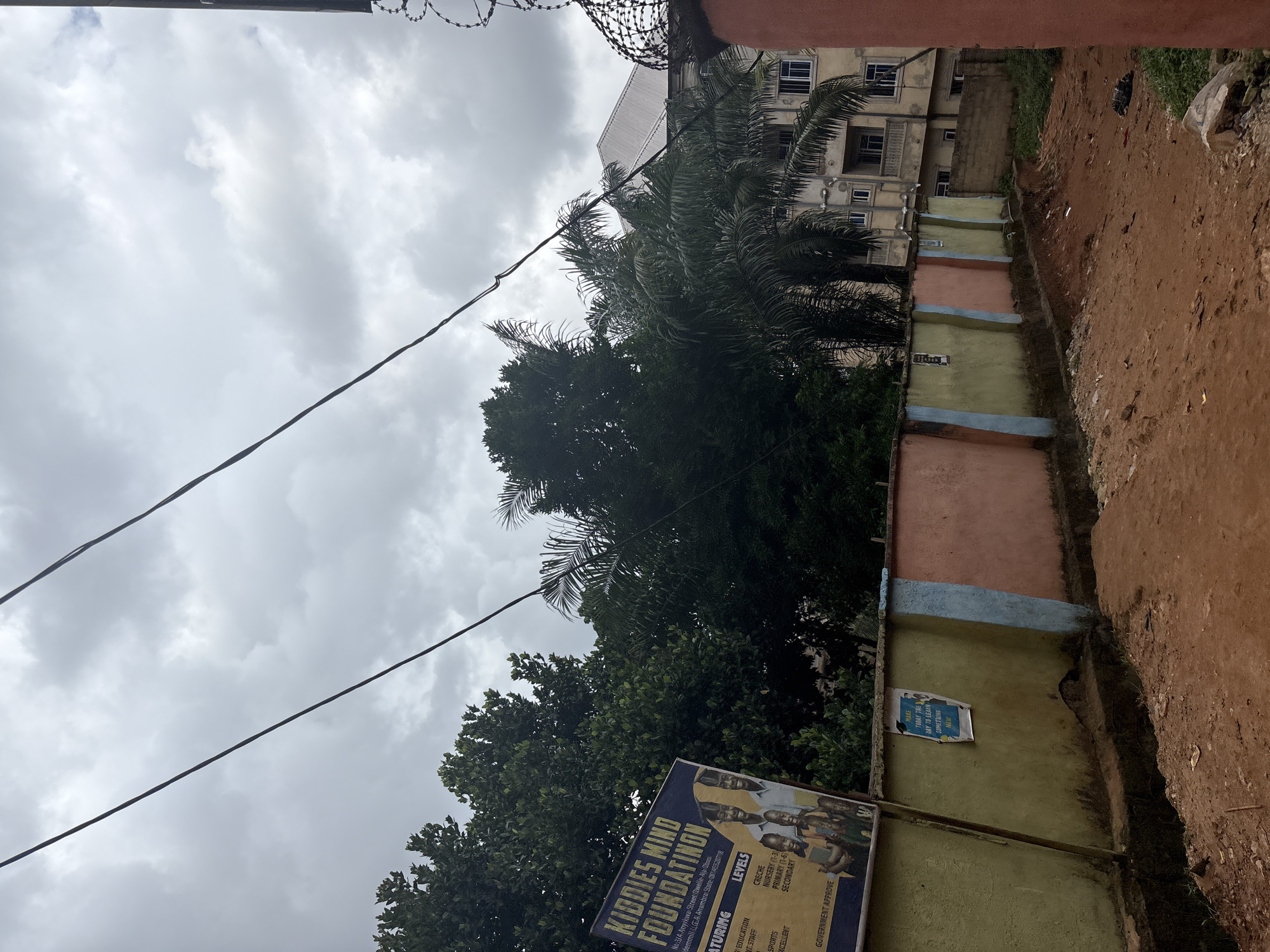 This is a medium shot, taken outdoors and oriented vertically, showing a cloudy sky above a building and trees. In the foreground, there are two power lines stretching across the frame. Below the sky and power lines, a multi-story building with visible windows stands tall. Palm trees and other lush green foliage surround the building.
A small, colorful poster or sign is affixed to a low wall in the middle ground, featuring images of children and text. The text on the sign is partially visible and includes phrases like "KIDDIES MIND FOUNDATION," "LEVELS," and "MAKE TODAY THE DAY TO LEARN SOMETHING NEW." The wall itself is painted in alternating bands of pastel colors like peach, light blue, and yellow-green.
The ground to the right of the wall appears to be reddish-brown dirt or mud, with some scattered debris. The overall scene suggests an urban or semi-urban environment in Nkpor, Nigeria, under a potentially overcast or approaching-rain sky. The presence of the foundation sign implies an educational or community-focused establishment.