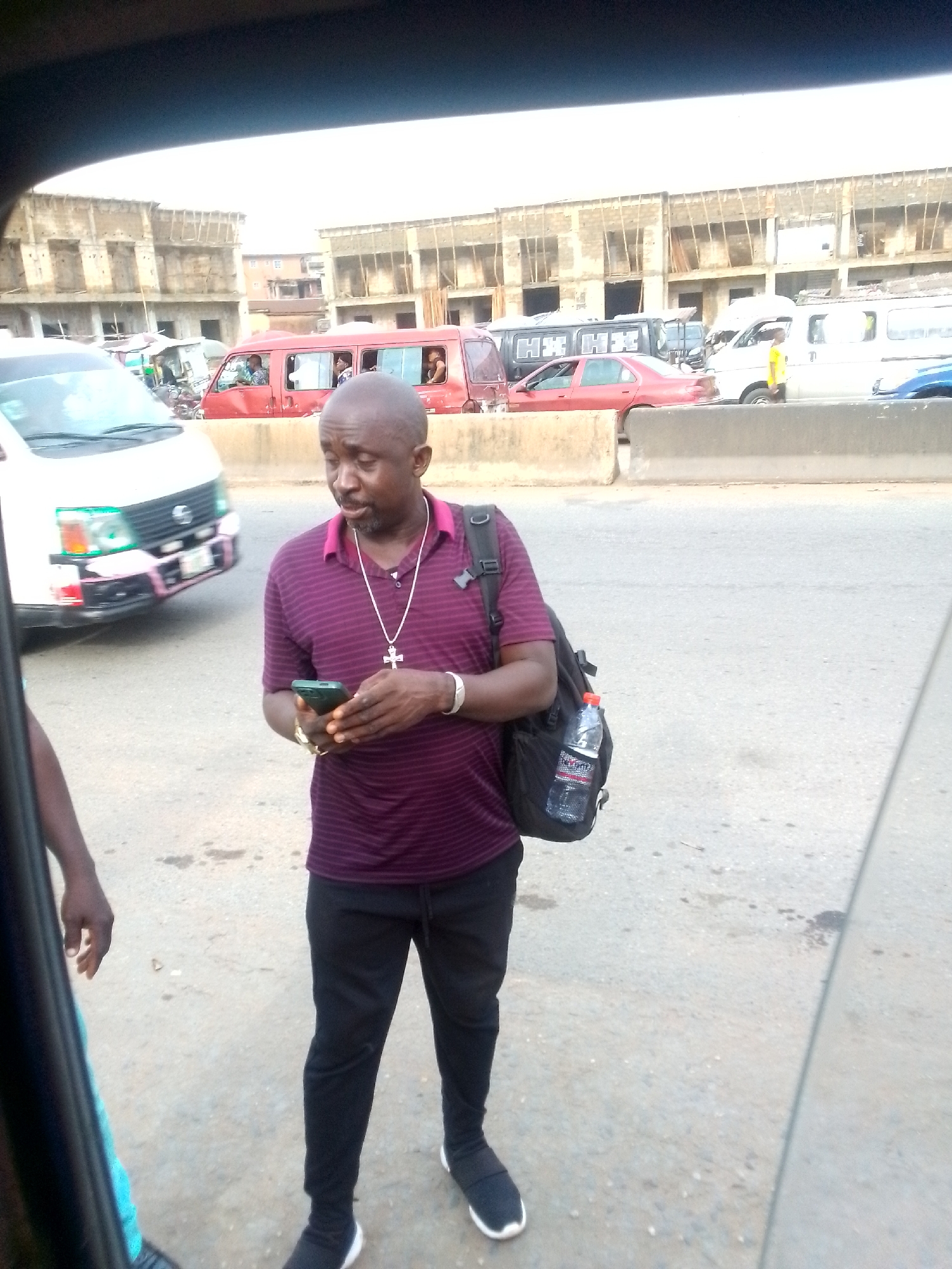 A man is standing on a street in Ihiala, Nigeria. He is holding a smartphone and is wearing a purple striped shirt, black pants, and black shoes. He has a backpack and a cross necklace. A white van and other vehicles are visible on the street behind him. Buildings are visible in the background.