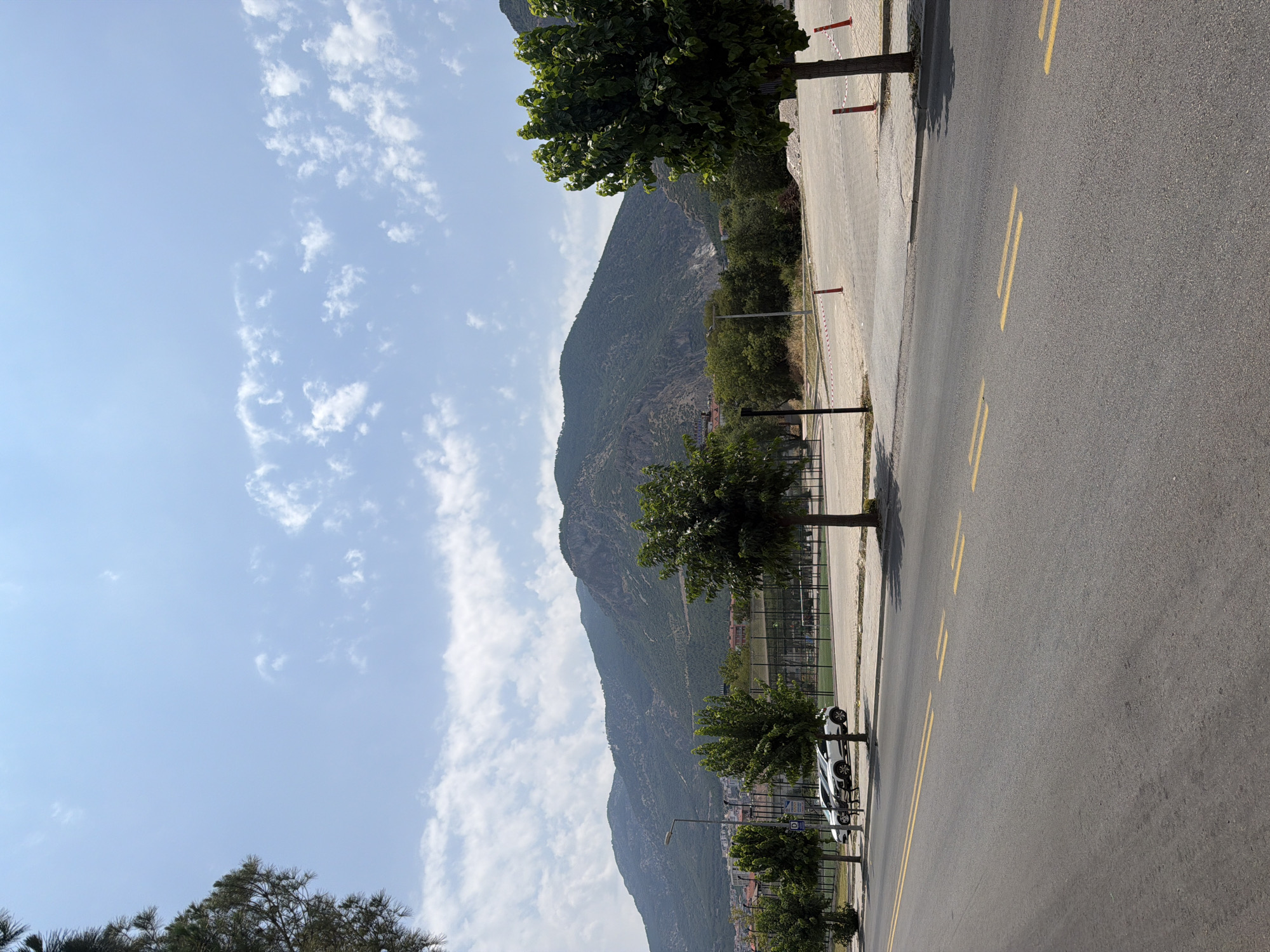 A landscape view shows a paved road in the foreground with yellow double lines marking lanes. To the left, a few trees line the road. Beyond the road, a fenced sports court with green turf is visible. In the background, a mountain range dominates the horizon under a blue sky with scattered white clouds. Several white cars are parked on the side of the road. The scene depicts an outdoor environment with elements of urban infrastructure and natural landscape. The overall impression is one of a sunny day in a town located near a mountainous region.