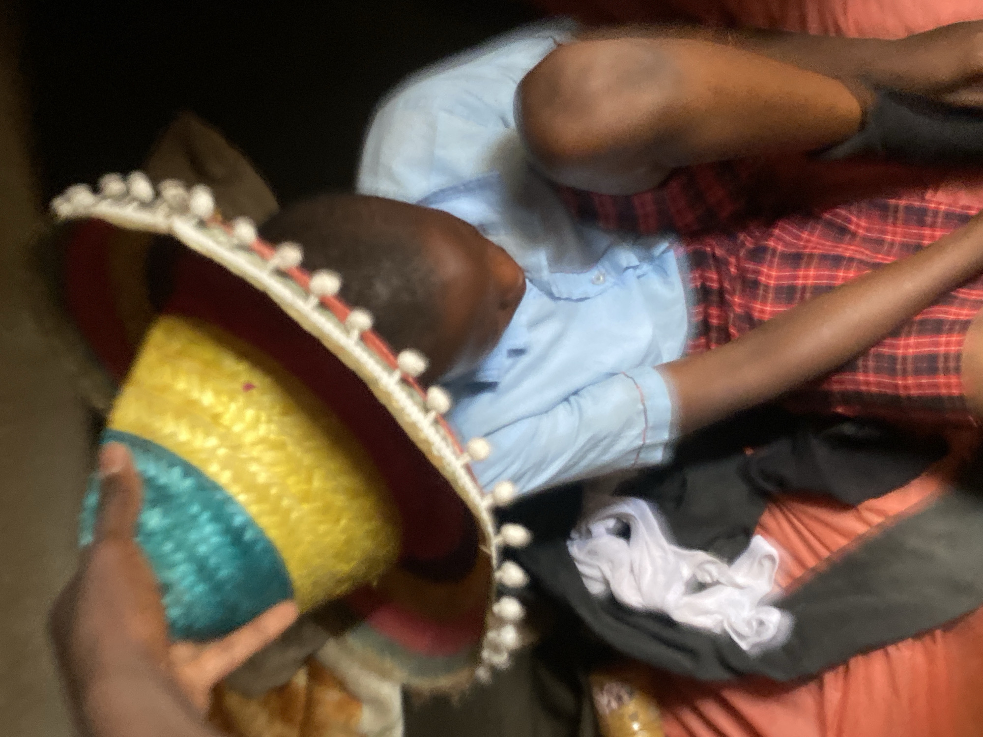 A person is lying on a bed with a plaid blanket and a light blue shirt. A hand holds a colorful sombrero. The location is purported to be Bukuru, Nigeria.