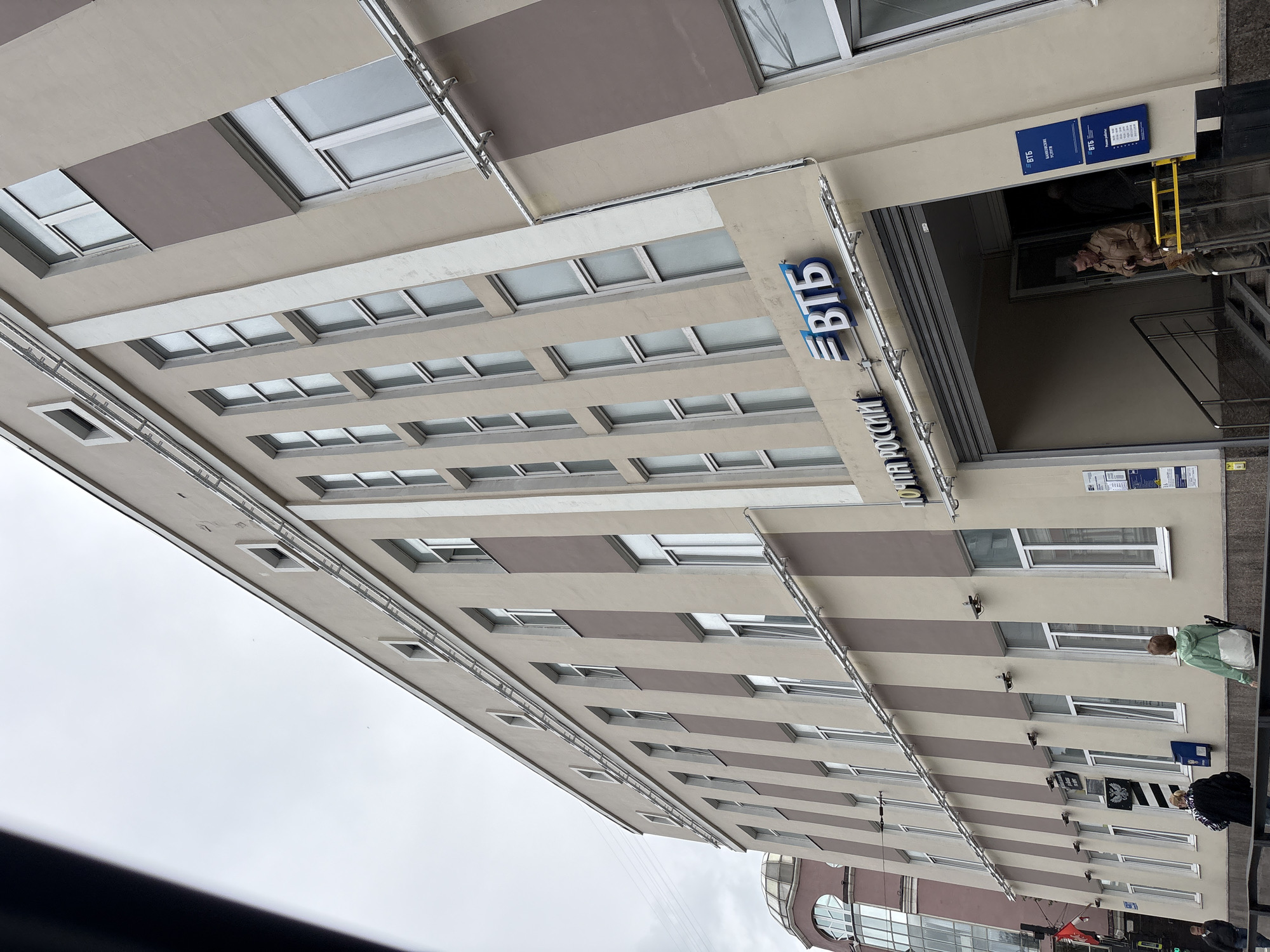 The image shows a multi-story building with a light beige facade and vertical brown stripes. The building has numerous windows, many of which appear to be closed. Above the entrance to the building, there are signs with Russian text. One sign reads "ПОЧТА РОССИИ" (Russian Post), and another sign displays the logo and name of "ВТБ" (VTB Bank). Below the VTB sign, there are blue signs indicating "БАНКОВСКИЕ УСЛУГИ" (Banking Services).
The entrance area features a ramp and stairs leading down into the building. A man in a tan jacket is walking down the stairs towards the entrance. Two other people, one in a black jacket with a white pattern and the other in a light green jacket, are walking away from the building on the sidewalk. There is a blue post box on the sidewalk near the people. A red flag is visible on a pole to the left of the building. In the background, another building with a domed roof is partially visible. The sky is overcast.
This building is located at Komsomolsky Prospekt, 66, Perm, Russia, which houses a VTB bank branch and a Russian Post office.