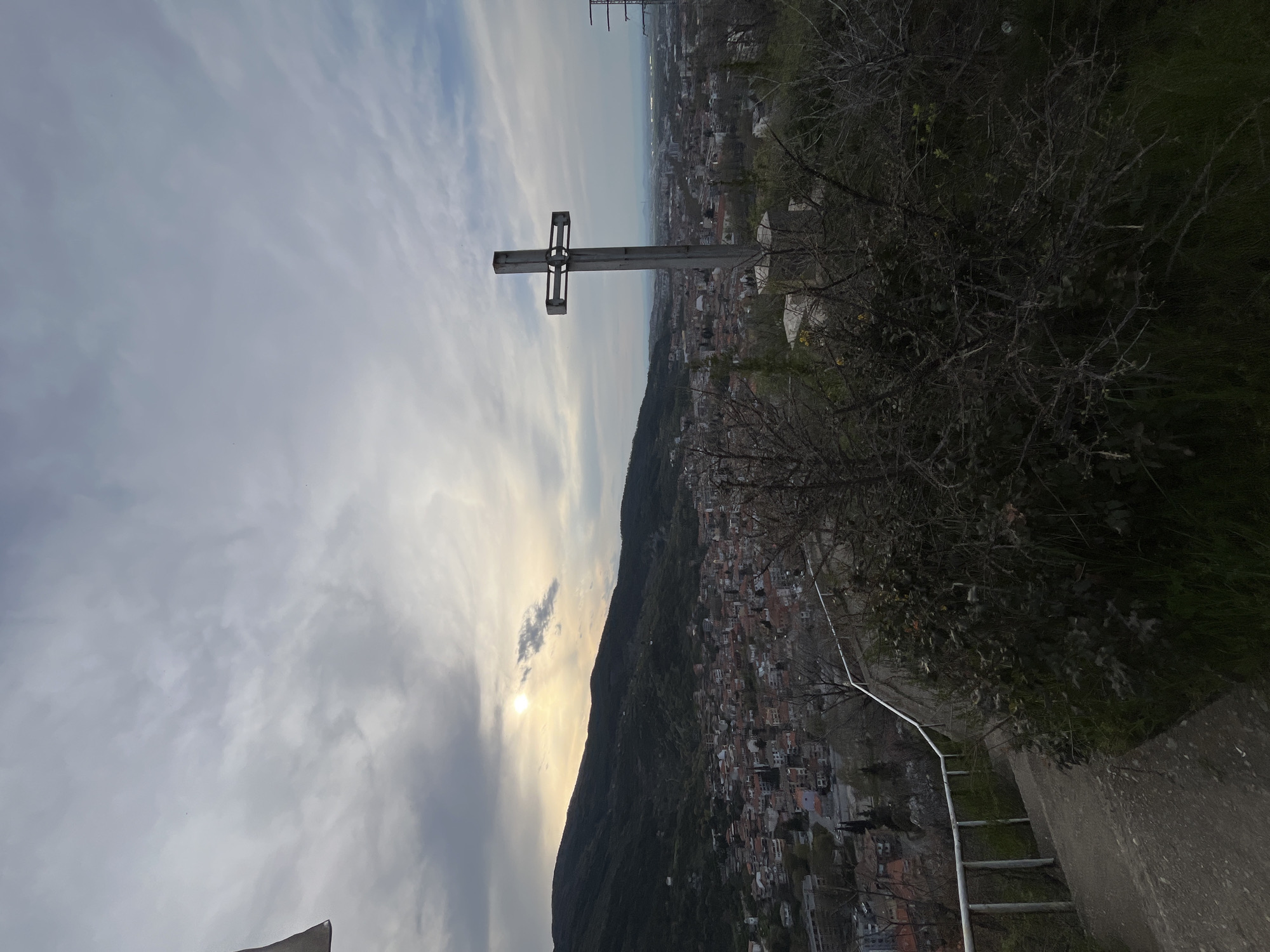 The image shows a metal Christian cross atop a hill overlooking the city of Asenovgrad, Bulgaria. The sky is filled with clouds and the sun is visible. A hillside with trees and vegetation is on the right. A metal railing and steps are visible on the right edge of the frame. The city of Asenovgrad stretches out below the hill, with buildings visible in the distance.