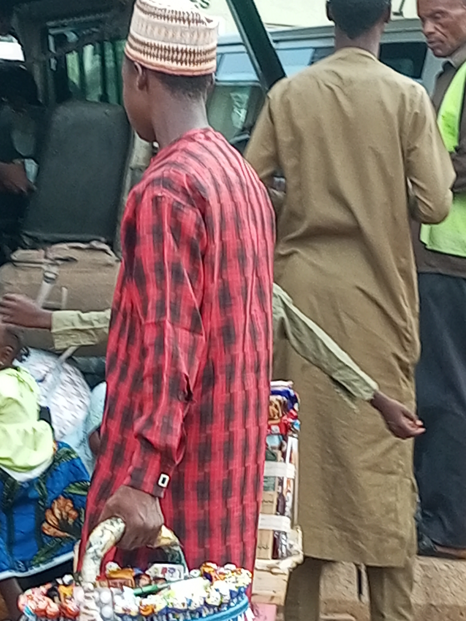 A man wearing a red and black checkered long-sleeved shirt and a white and brown patterned cap stands in the foreground, facing away from the camera and holding a basket of goods. The basket contains various small items and appears to be a form of street vending. Behind him, another man in a tan tunic is visible, also facing away. In the background, a vehicle is partially seen, suggesting an outdoor, possibly urban, setting. The overall impression is of a busy market or roadside scene.