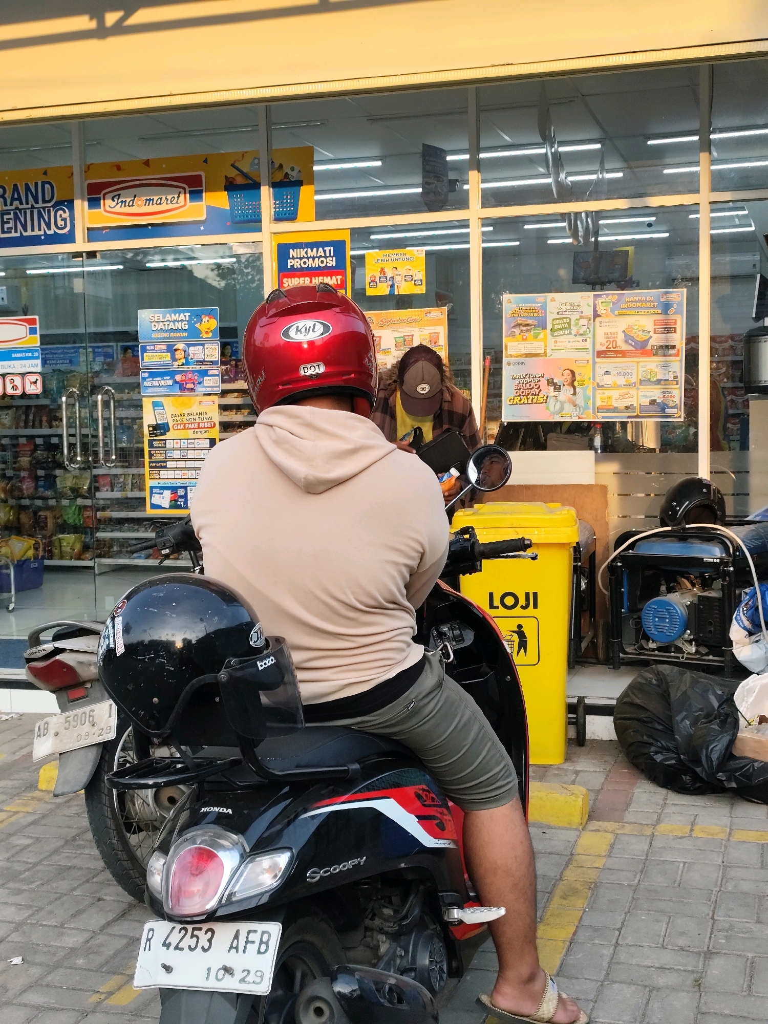 The image captures a scene outside an Indomaret convenience store in Sanden, Indonesia. The foreground is dominated by the back of a person sitting on a black scooter, wearing a beige hoodie and olive-green shorts. A red helmet with "KYT" branding is on their head, and a black helmet is secured to the back of the scooter. The scooter's license plate reads "R 4253 AFB". To the left, another scooter's rear is partially visible, with a license plate reading "AB 5906 I".
Behind the scooter, a yellow trash bin with the word "LOJI" is prominent. To the right of the bin, a blue and silver generator is visible, partially covered by a large black garbage bag. The store's entrance is visible, with glass doors and various promotional posters displayed inside and outside. One poster advertises "GRAND OPENING" and "INDOMARET". Another poster highlights "NIKMATI PROMOSI SUPER HEMAT". Inside the store, shelves stocked with products can be seen. The ground is paved with grey and yellow checkered tiles. The lighting suggests it is late afternoon, with long shadows cast on the ground. A person is visible inside the store behind the counter, wearing a brown shirt and a baseball cap. The overall impression is a casual stop at a local convenience store.