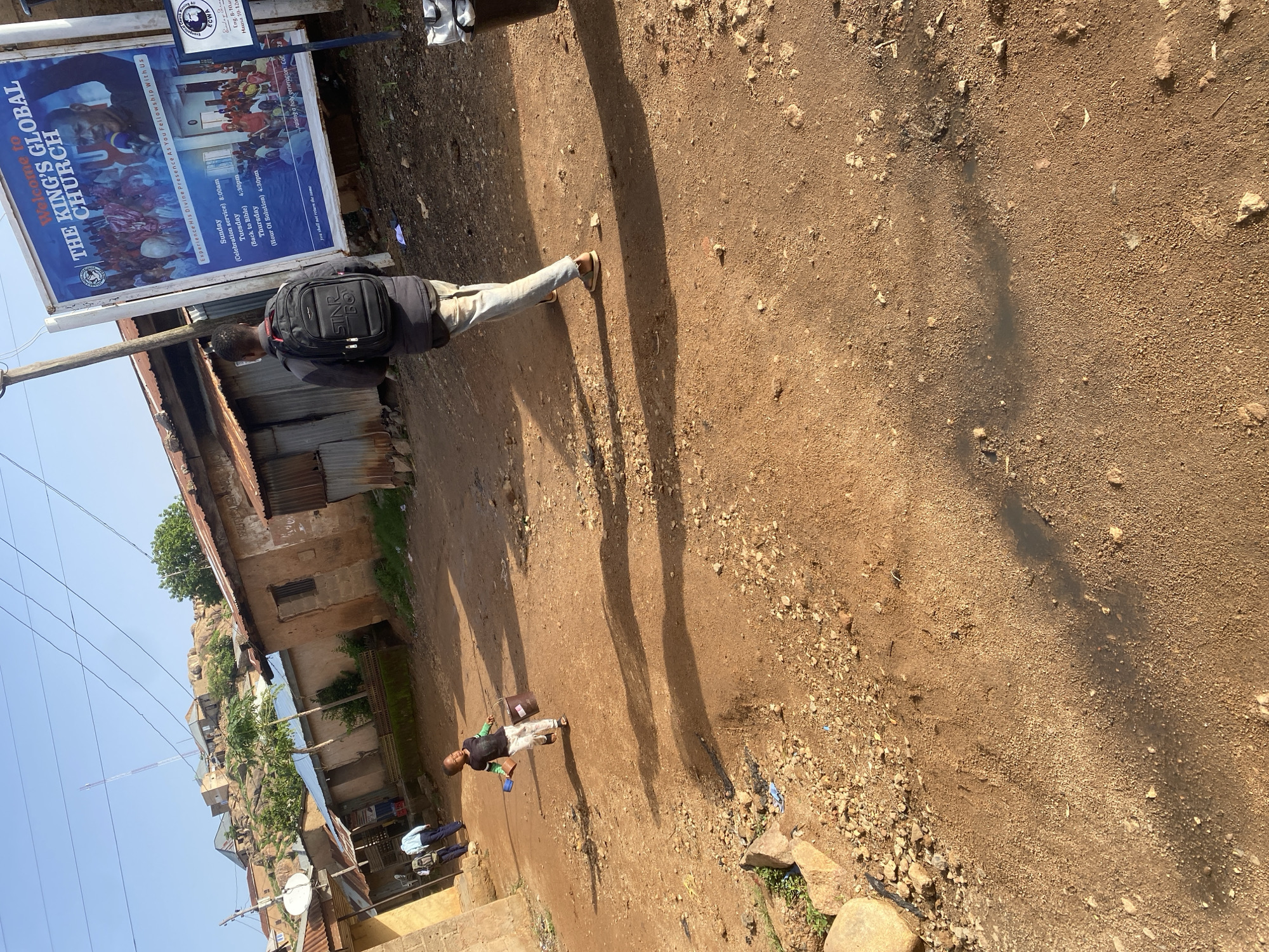 The image depicts a street scene in Jos, Nigeria, under a clear blue sky. A young child, wearing a black long-sleeved shirt and white pants, is walking away from the camera carrying two buckets. To the right, a man with a black backpack walks towards the camera. Behind him, a large billboard advertises "THE KING'S GLOBAL CHURCH" with service times and images of church activities. The street is unpaved and dusty, with scattered rocks and debris. On either side of the street, buildings made of earth and corrugated iron are visible, some appearing weathered and incomplete. In the background, more structures are perched on a rocky hillside, characteristic of parts of Jos. The overall impression is of a typical urban environment in Nigeria, characterized by its unique architecture and the presence of both religious and residential elements.
