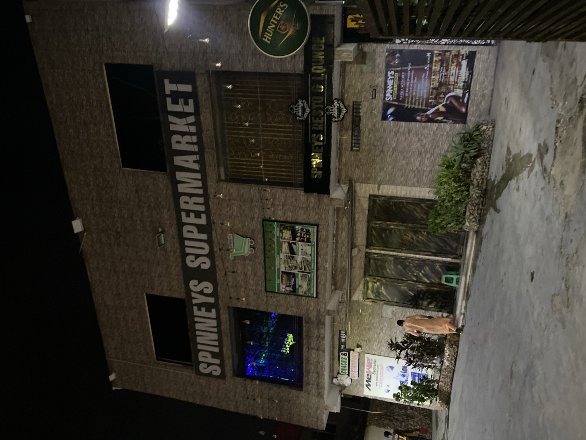 The image shows the exterior of a Spinneys Supermarket and Spinneys Resto & Lounge in Ikorodu, Nigeria. The building is multi-storied with brick facade. The signage "SPINNEYS SUPERMARKET" is vertically placed on the left side. Other signs include "HUNTER'S," "SNACKS & GRILL," and "SPINNEYS RESTO & LOUNGE". A promotional banner for "MEWE Electronics" is visible at the bottom. A person is seen walking towards the entrance. The environment seems to be during night.