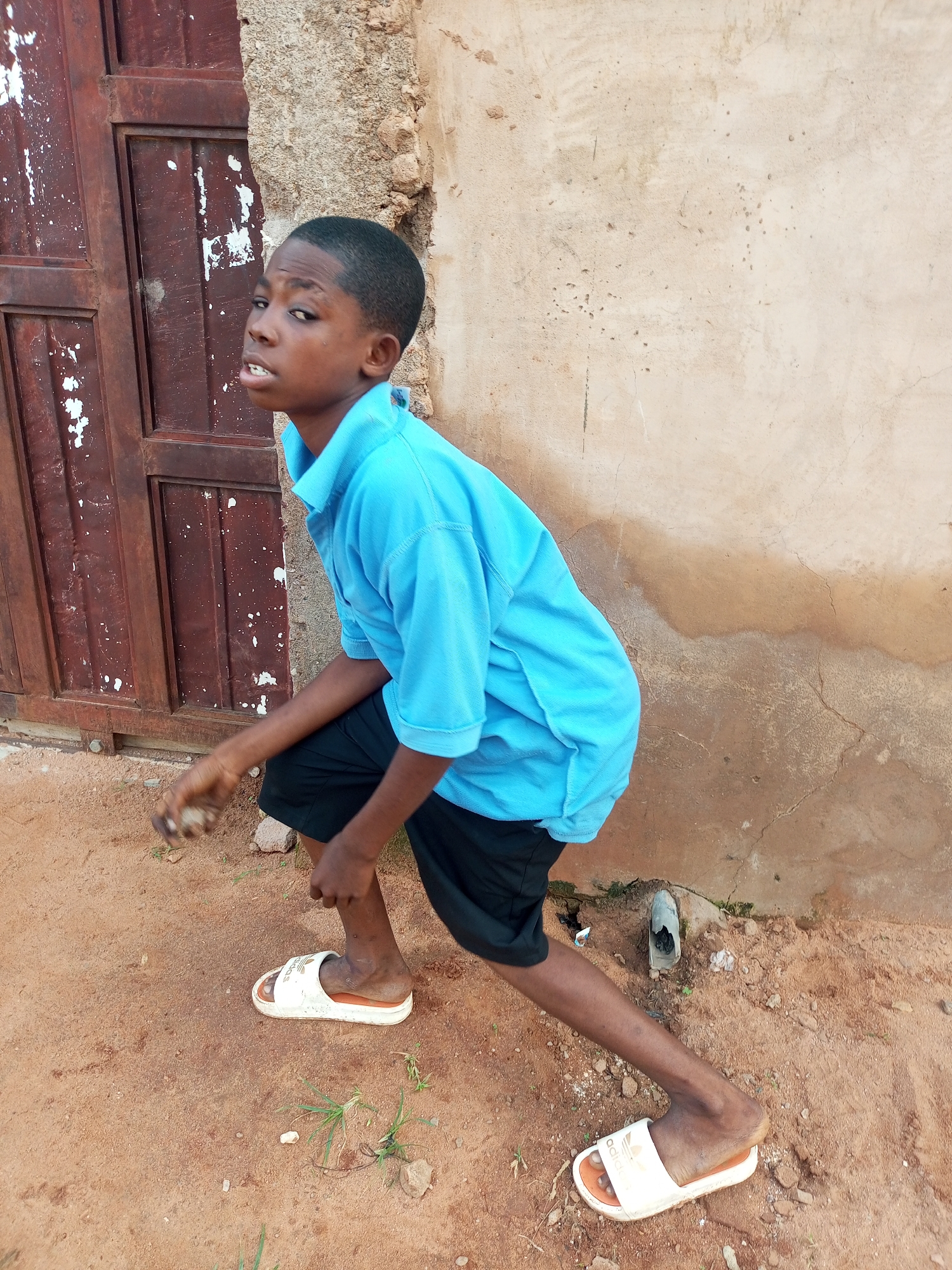 In Tempure, Nigeria, a young boy stands in front of a weathered, dark brown wooden door with white paint splatters. The boy, wearing a blue polo shirt and black shorts, is positioned on reddish-brown dirt ground. He is looking to his left with an open-mouthed expression. His right hand is held slightly forward, appearing to be holding a small object. He is wearing white slide sandals. Behind him, a rough, light-colored stucco wall shows signs of age and discoloration. The ground is a mixture of sand and small stones, with sparse green vegetation. To the right of the boy, near the wall, a small, broken pipe protrudes from the ground.