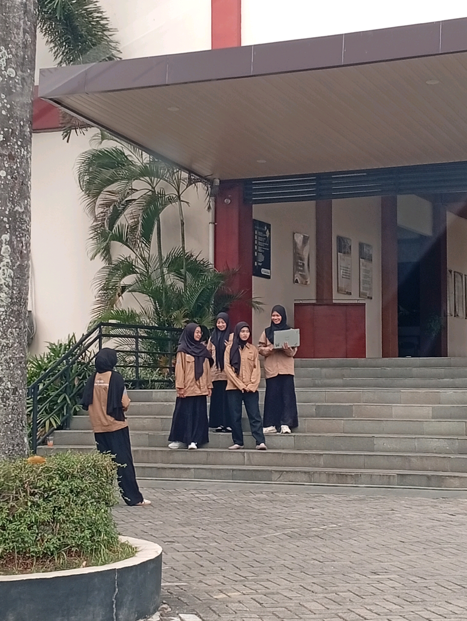 Six individuals, four women and two others not clearly identifiable as male or female, stand on the steps of a building with a light-colored facade and a dark awning. The women are all wearing light brown jackets and dark skirts or pants, and headscarves. The woman second from the right is holding a silver laptop. The building appears to be an educational institution, possibly a university, given the presence of posters on the wall. Palm trees and other foliage are visible around the building. The ground in front of the steps is paved with interlocking bricks, and a low, rounded planter with green bushes is on the left side of the image. The scene takes place outdoors during daylight hours.