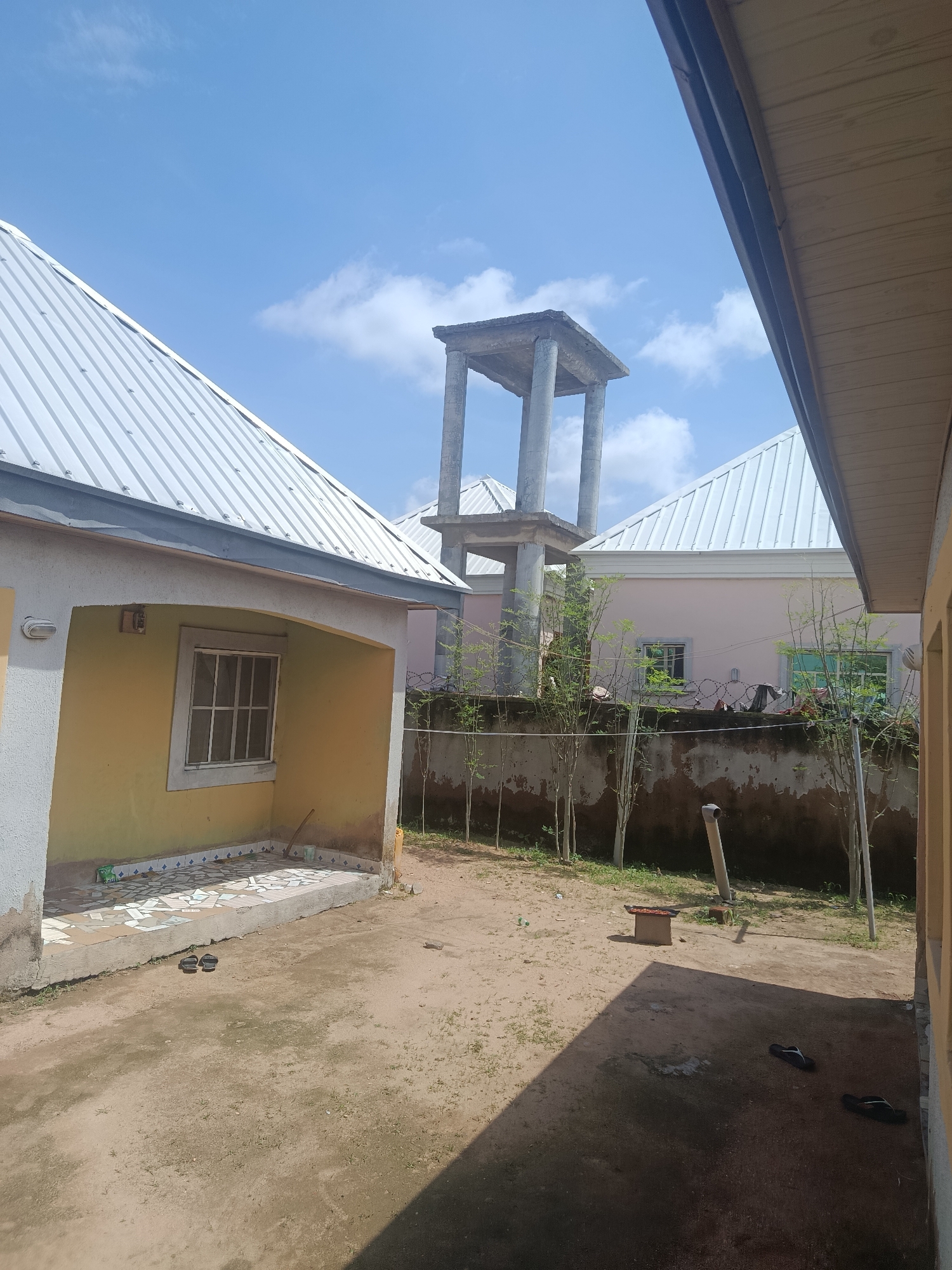 The image captures an outdoor courtyard with two houses and a water tower under a bright, partly cloudy sky. The foreground is dominated by a sandy, uneven ground with scattered patches of dry grass. Two pairs of dark flip-flops are visible on the ground, one near the foreground house and another further back in the shade.
To the left, a yellow house with a white arched entrance features a tiled patio and a window with a metal grate. A light fixture is mounted on the exterior wall above the entrance. To the right, a portion of another building, possibly a veranda with a wooden soffit, is visible.
In the background, a light pink house with white trim and a white metal roof is seen. This house has two windows with reflective glass. A low concrete wall separates the courtyard from the pink house, topped with barbed wire fencing. Young trees with sparse foliage are planted in a line along the wall.
A concrete water tower with four pillars supporting a platform stands in the mid-ground, between the two houses. A small, square metal object, possibly a cooking grill, rests on the ground in the middle distance, casting a shadow. A plastic bottle and a section of white pipe are also visible on the ground. The scene suggests a residential area in Jauro Yinu, Nigeria, possibly a compound with ongoing construction or recent development, given the unfinished look of the water tower.