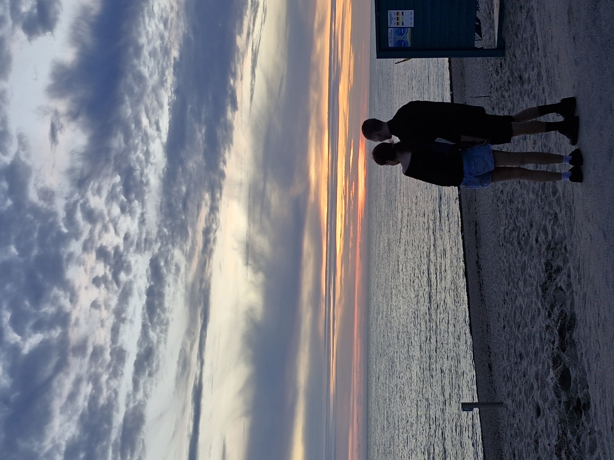 A couple stands on a sandy beach in Klaipėda, Lithuania, facing the sea at dusk. The sky is filled with dramatic clouds, painted with hues of orange, pink, and blue, indicating the setting sun. The sea is calm, with gentle waves lapping the shore. A blue lifeguard stand is visible to the right of the frame. The couple, silhouetted against the colorful sky, appear to be embracing or standing close together, enjoying the view. The overall scene evokes a sense of peacefulness and romanticism.