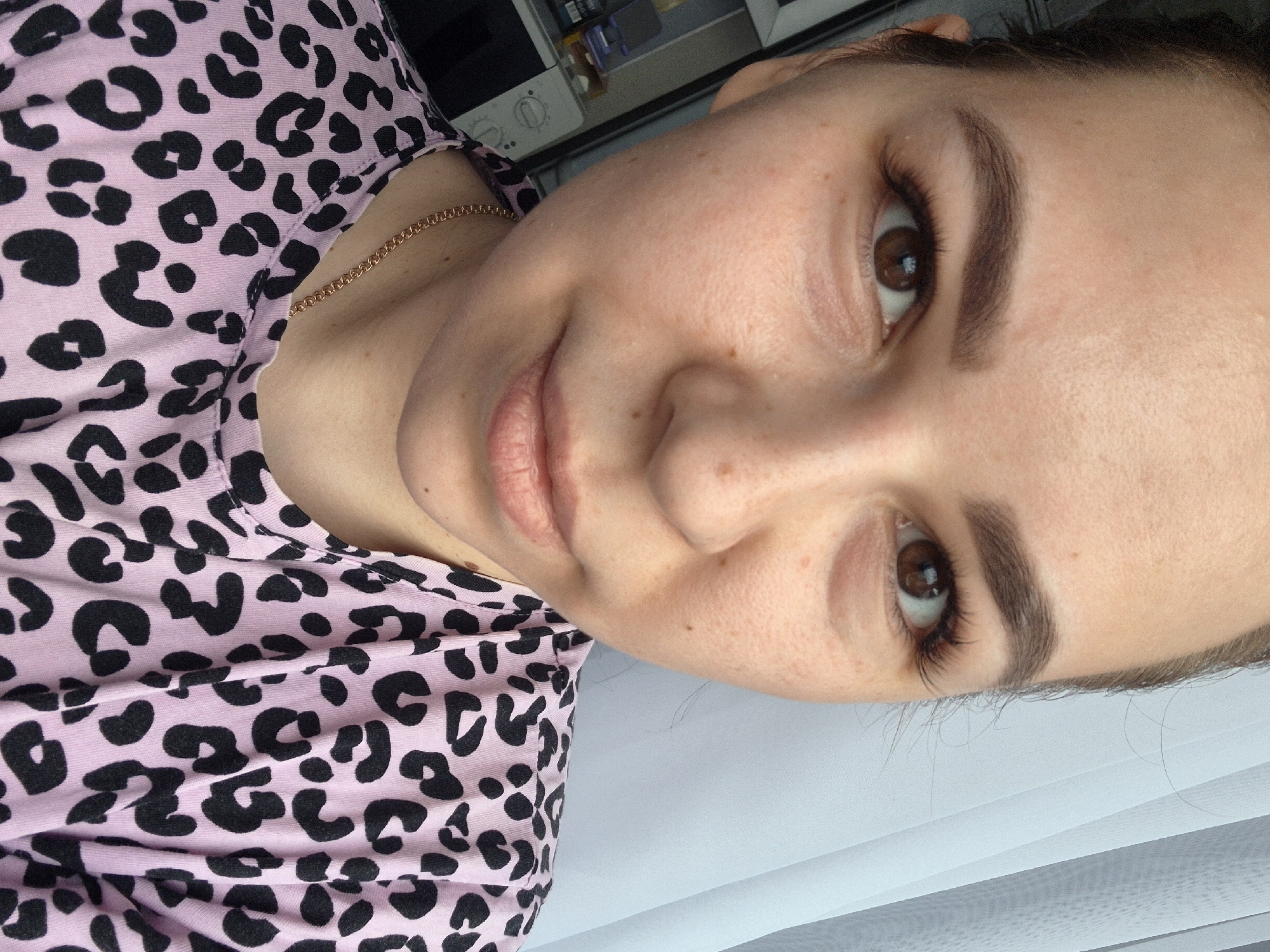 A close-up portrait of a woman with fair skin, brown eyes, and dark eyebrows. She is wearing a pink leopard print top and a thin gold chain around her neck. The background is blurry, but appears to show a window with closed blinds. There is no information to determine that the location is in the city of Tobolsk, Russia.