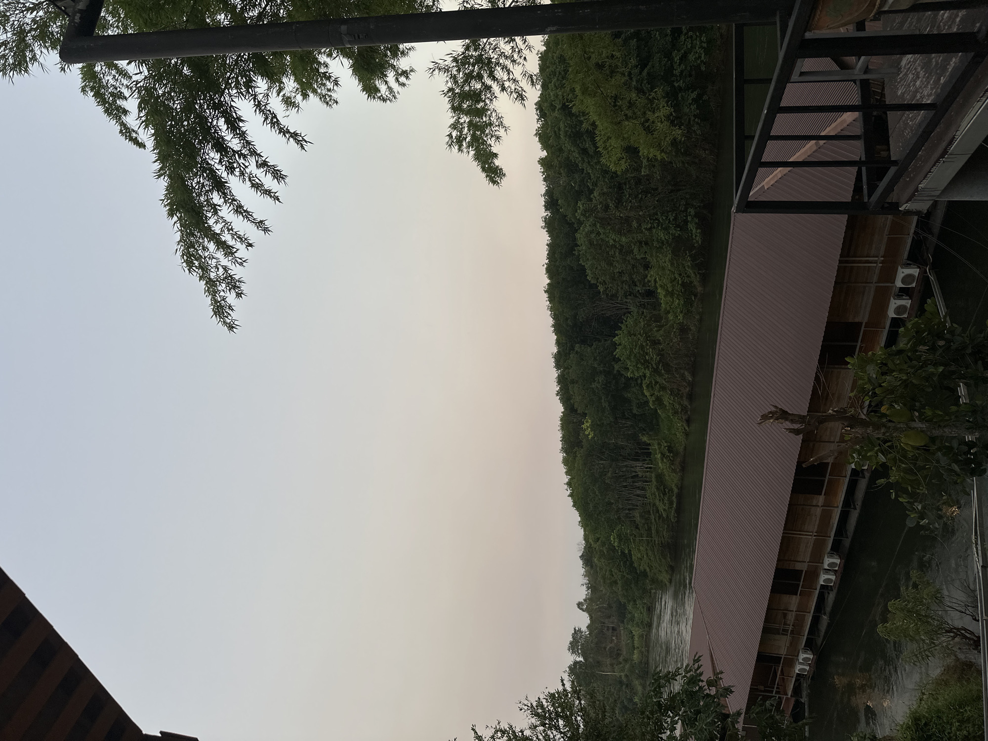 The image shows a waterscape in Sai Yok, Thailand, at what appears to be dusk. The sky is a uniform light color, likely due to overcast conditions or haze. A densely forested area borders a body of water, possibly a river, that stretches into the distance. On the right side of the image, a structure with a reddish-brown roof is visible, possibly a resort or dwelling built over the water. Air conditioning units are visible on the exterior of this structure. Green foliage is present both in the foreground and hanging from above. A black metal structure, possibly a railing, is also partially visible at the top-right.