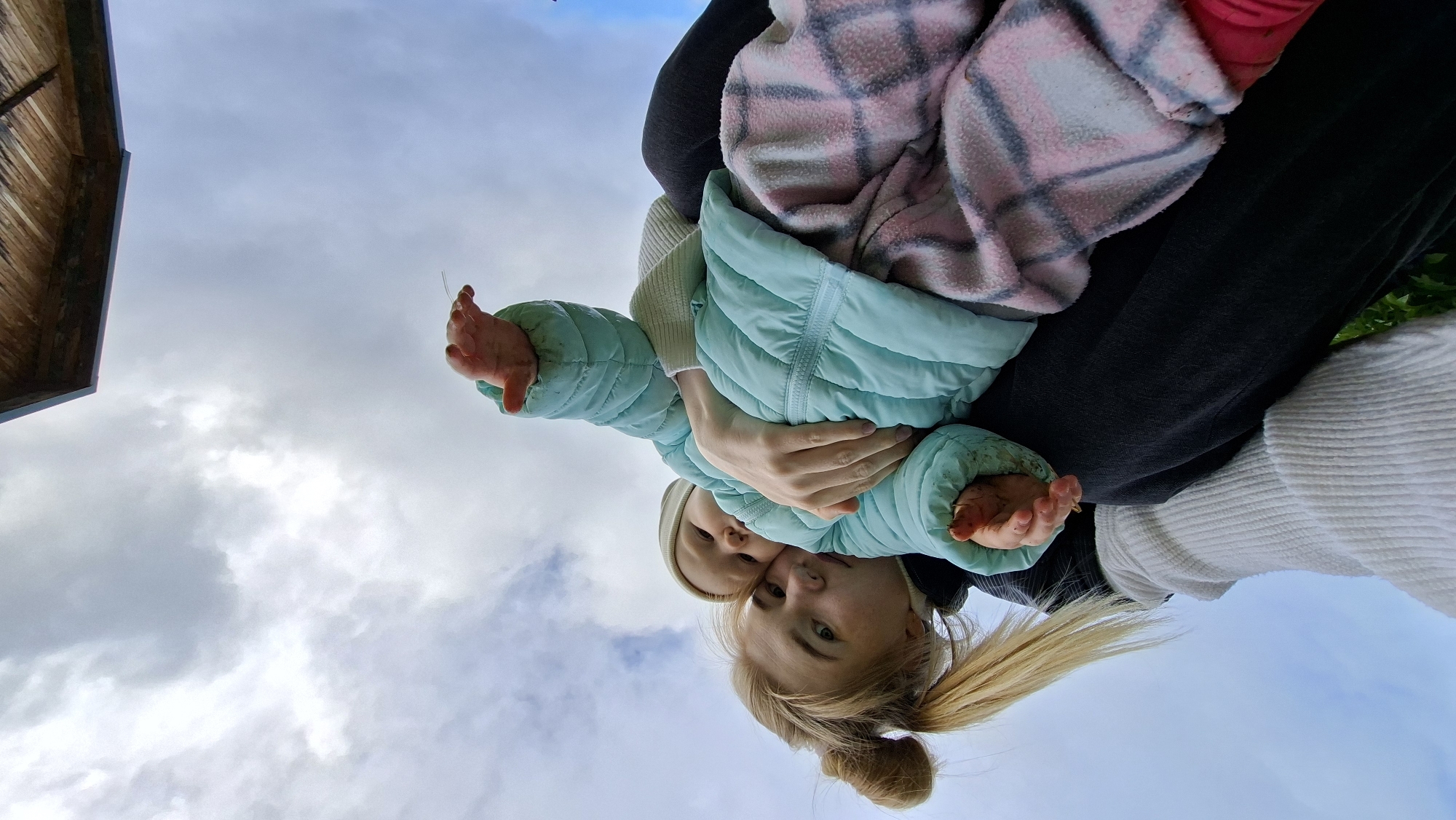 From a low angle, a woman with blonde hair in a high bun holds a baby in a light blue jacket. The baby also wears a light-colored hat. Both are facing the camera. Above them, there's a cloudy sky and the edge of a wooden structure. A pink and grey blanket can be seen on a person's lap. The location is Большой Букор, Russia.