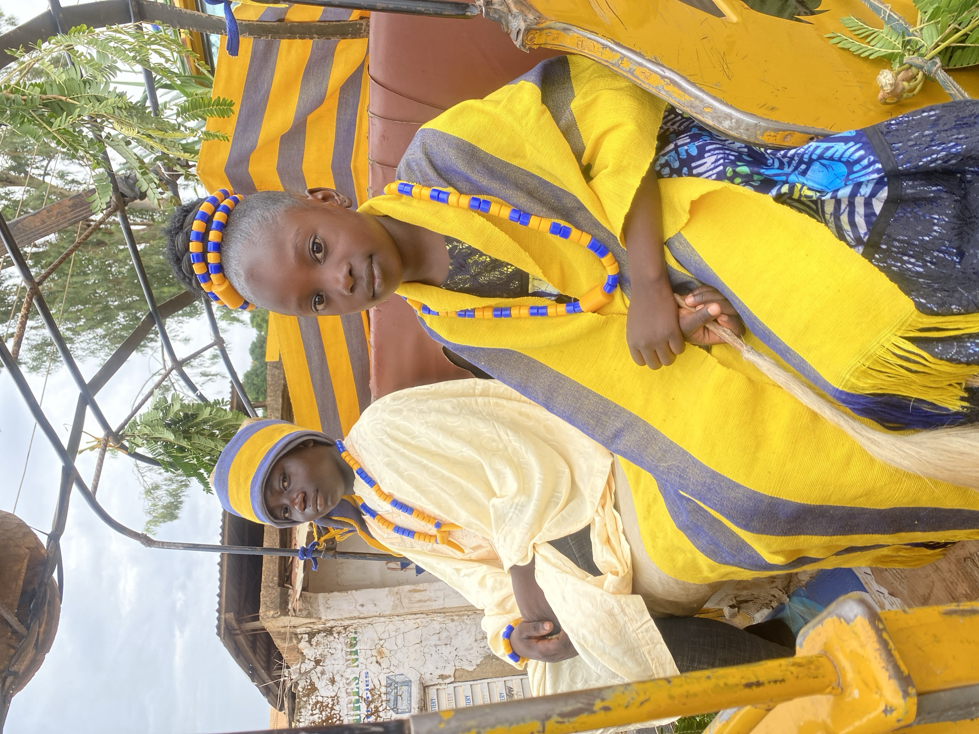 Two individuals are photographed indoors, likely within a vehicle or structure associated with transportation. The younger individual, positioned in the upper part of the frame, is adorned with yellow and blue beaded jewelry around their head and neck. They are draped in a yellow shawl with blue stripes. Below, the other individual wears a matching hat and neckwear, also in yellow and blue.
The surrounding environment includes vegetation visible through a frame. An exterior view reveals a building structure in the background, partially obscured by the frame, suggesting an urban setting, plausibly Maigizo, Nigeria. The overall impression is one of cultural attire and a posed shot within an identifiable location.