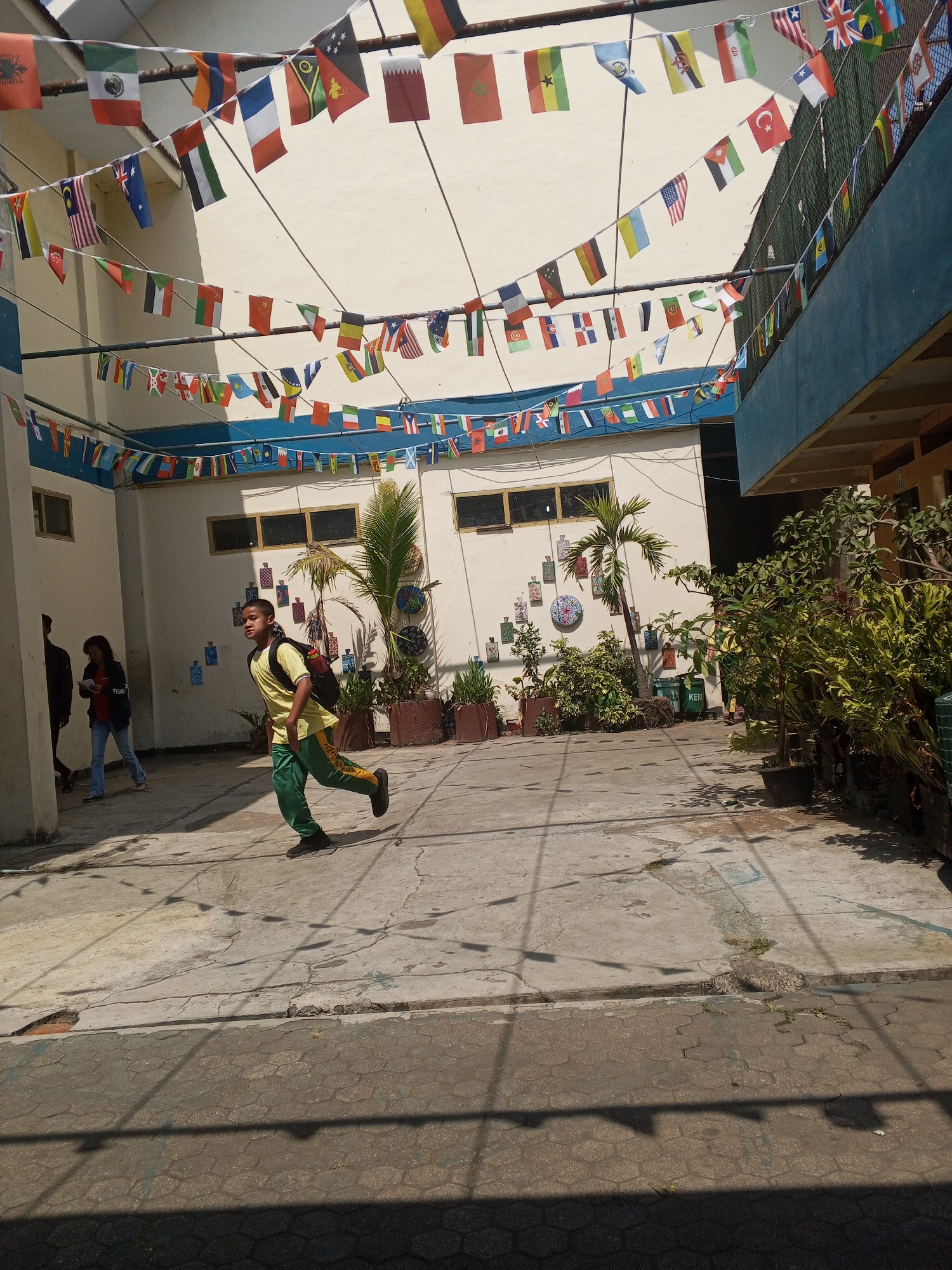 The image shows an outdoor courtyard at what appears to be a school or community center in Turen, Indonesia, as suggested by context. The courtyard has a paved floor with some cracks and a bordering area of hexagonal paving stones. Numerous strings of international flags are suspended overhead, creating a festive atmosphere. In the middle ground, a young person wearing a yellow t-shirt and green pants with yellow stripes is running across the courtyard, carrying a backpack. To the left, two other individuals are walking away from the camera. The background features white walls of buildings with windows, some potted plants, and a palm tree. Sunlight casts shadows on the ground, indicating it is daytime.