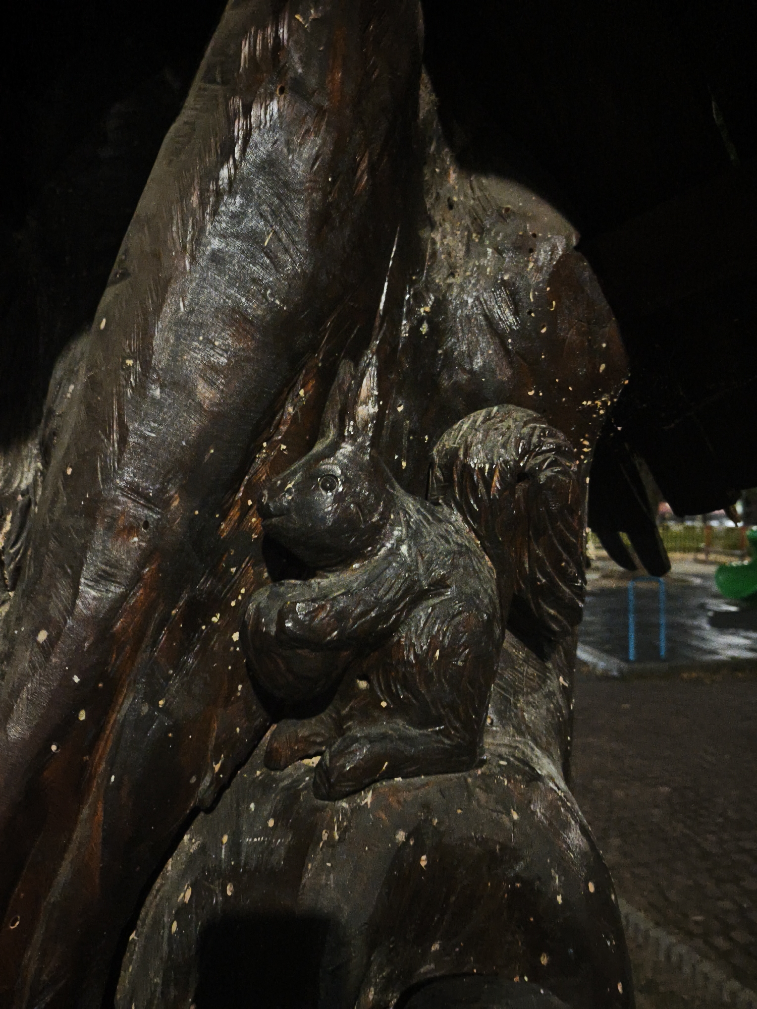 The image shows a close-up, low-angle view of a wooden sculpture of a squirrel resting on a tree trunk. The squirrel is carved in a dark brown wood and is depicted with its head turned to the left, its paws resting on its chest, and its bushy tail curled upwards. The tree trunk sculpture is textured with horizontal lines suggesting bark. Small light-colored specks, possibly from leaves or debris, are scattered across the surface of the sculpture. In the background, out of focus, there is a dark area with some blurry shapes of playground equipment, including blue poles and a green slide, suggesting the sculpture is located in an outdoor public area like a park or playground. The lighting is dim, creating a dramatic effect and highlighting the texture of the wood. This sculpture is located in the city of Lovech, Bulgaria, specifically in an area commonly known as "Varosha" or near the "Kvartal Varosha" neighborhood, which is famous for its preserved traditional Bulgarian Revival architecture and its many wood carvings that adorn public spaces, often depicting local flora and fauna.