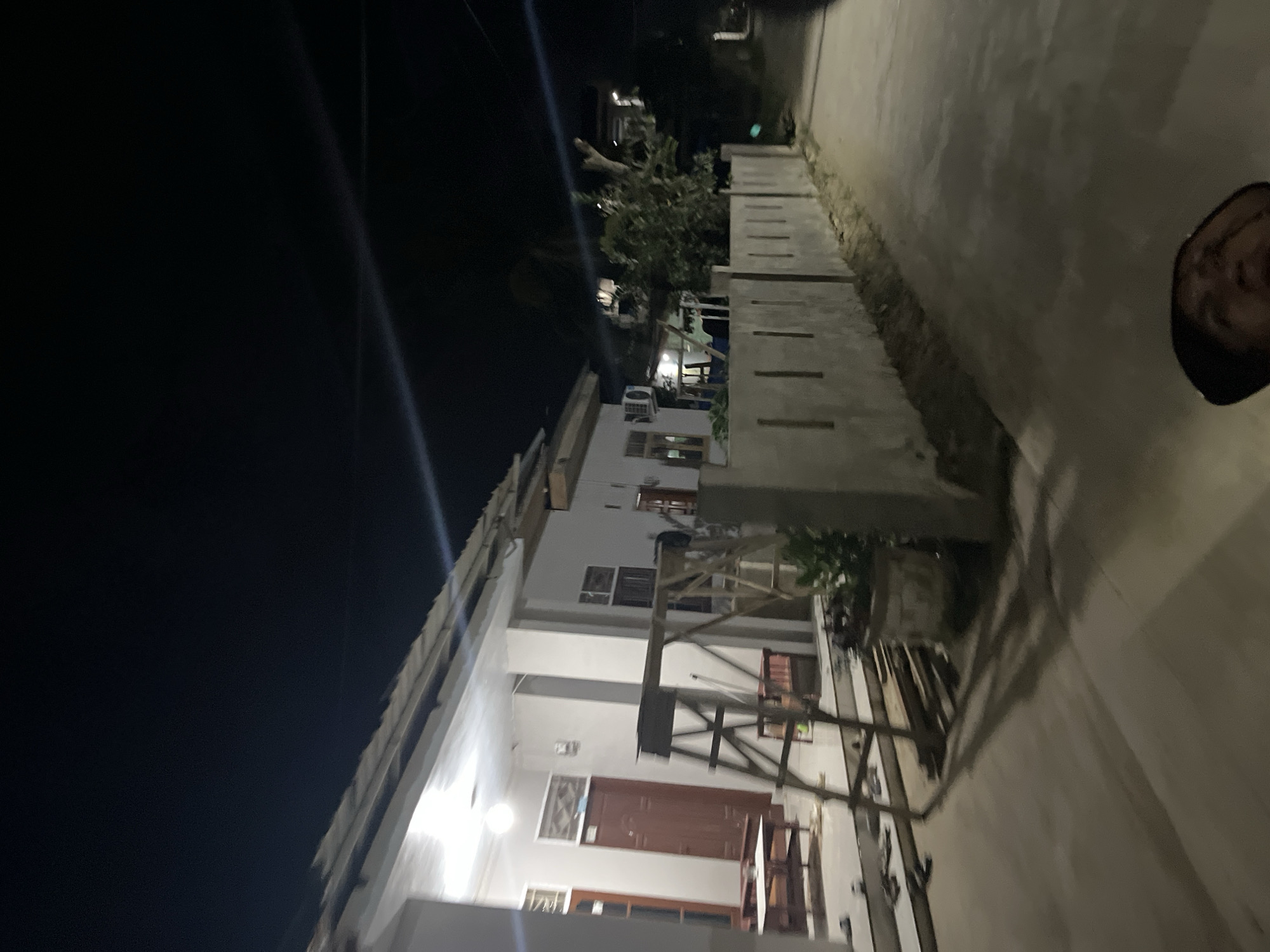 The media file depicts a nighttime scene in what appears to be a residential area. The primary subject is a multi-story building with a white facade, featuring several windows and doors. Construction scaffolding is visible in front of the building, suggesting ongoing work. An air conditioning unit is mounted on one of the walls. The building is illuminated by artificial lights, casting a glow on its exterior and the surrounding area.
To the right of the building, a pathway or walkway is visible, bordered by a low wall. In the background, trees and other structures can be partially seen, partially obscured by darkness. A sliver of a person's face is visible on the right edge of the frame, peeking out from behind the wall, with a smile visible.
The scene is set at night, as indicated by the dark sky and artificial lighting. There are no clear indications of weather conditions. The location is identified as Menanggini, Indonesia. The overall mood is somewhat utilitarian, possibly highlighting the ongoing construction or a typical evening in a residential neighborhood. No visible text is present in the image.