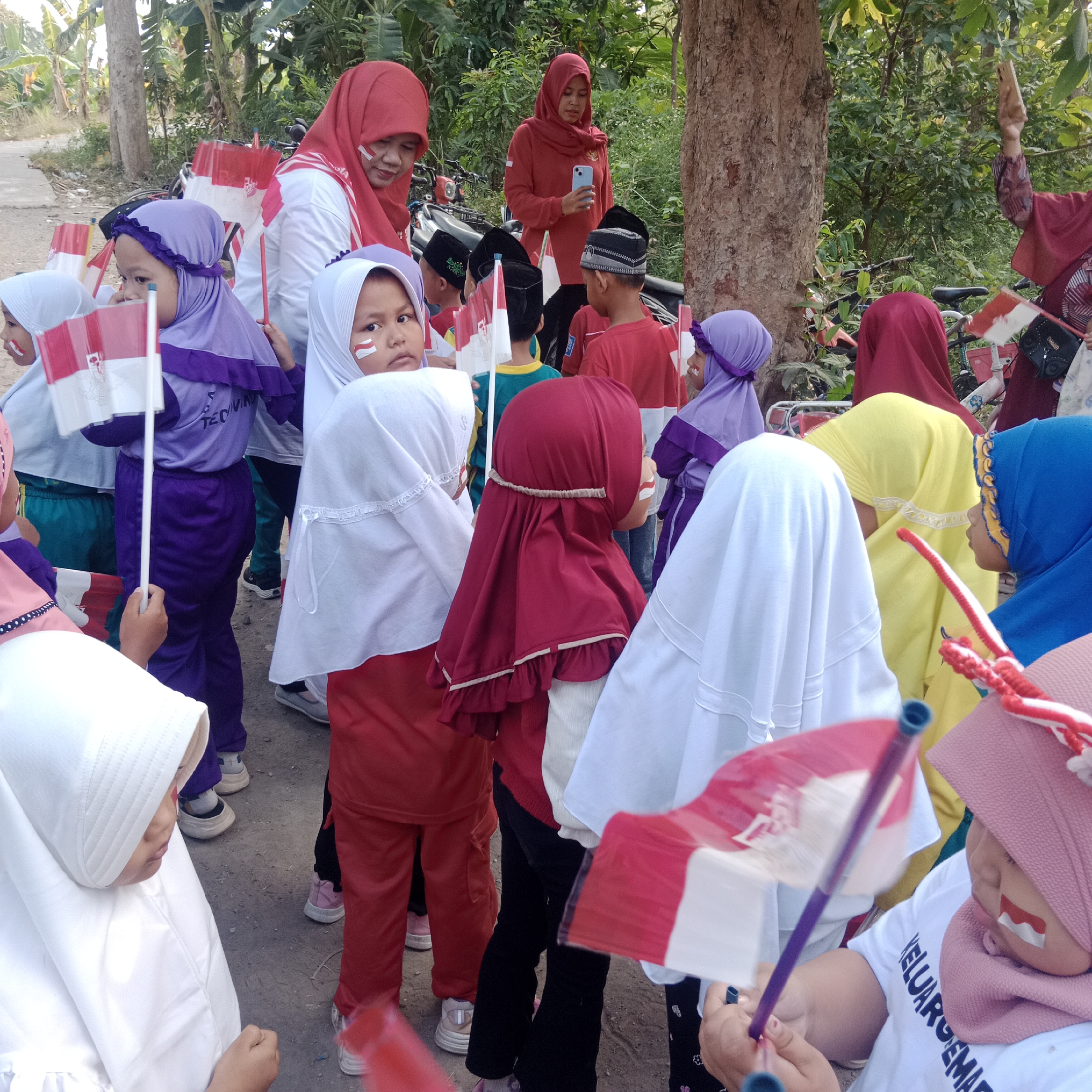 The image captures a group of children and adults in what appears to be an outdoor setting in Tegowanu Kulon, Indonesia, possibly during a celebration. Many of the individuals are wearing hijabs or headscarves and some children have red and white paint on their cheeks, resembling the Indonesian flag. Several people are holding small Indonesian flags.
In the foreground, a young girl wearing a white hijab and a pink headscarf is looking down, holding a flag. To her left, another child in a white hijab is visible. Behind them, a child in a purple hijab and matching pants holds an Indonesian flag. Further back, a woman in a red hijab and white top stands near a motorcycle, holding a smartphone. Another woman in a red top is also present. Other children in various colored hijabs and clothing are scattered throughout the scene, some holding flags. The background consists of trees and foliage, suggesting a rural or semi-rural environment. The ground is a dirt or gravel surface. The overall atmosphere appears festive and patriotic.