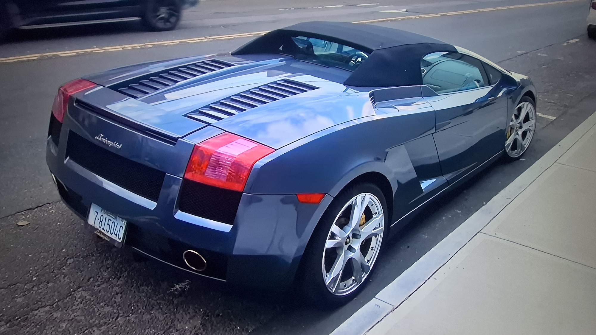 An overhead shot captures a dark blue metallic Lamborghini Gallardo Spyder convertible parked alongside a curb on a street. The car is oriented with its rear and right side visible, positioned next to a paved sidewalk. The black fabric convertible top is raised.
Notable features of the Lamborghini include its distinct louvered engine cover, large red taillights, and polished multi-spoke silver wheels with yellow brake calipers visible through the spokes. Two circular exhaust tips are present at the rear. The car's sleek body reflects the bright sky and clouds, suggesting a daytime setting with potentially overcast or partly cloudy weather.
In the background, a dark-colored vehicle is partially visible on the street, and double yellow lines mark the road. The car is parked on asphalt, which appears somewhat worn in places.
Visible text includes the brand name "Lamborghini" embossed on the rear of the vehicle, above the license plate. The license plate itself displays the characters "7-81504C". Although faint, the word "DELAWARE" is partially discernible above the numbers on the license plate, indicating its state of origin.