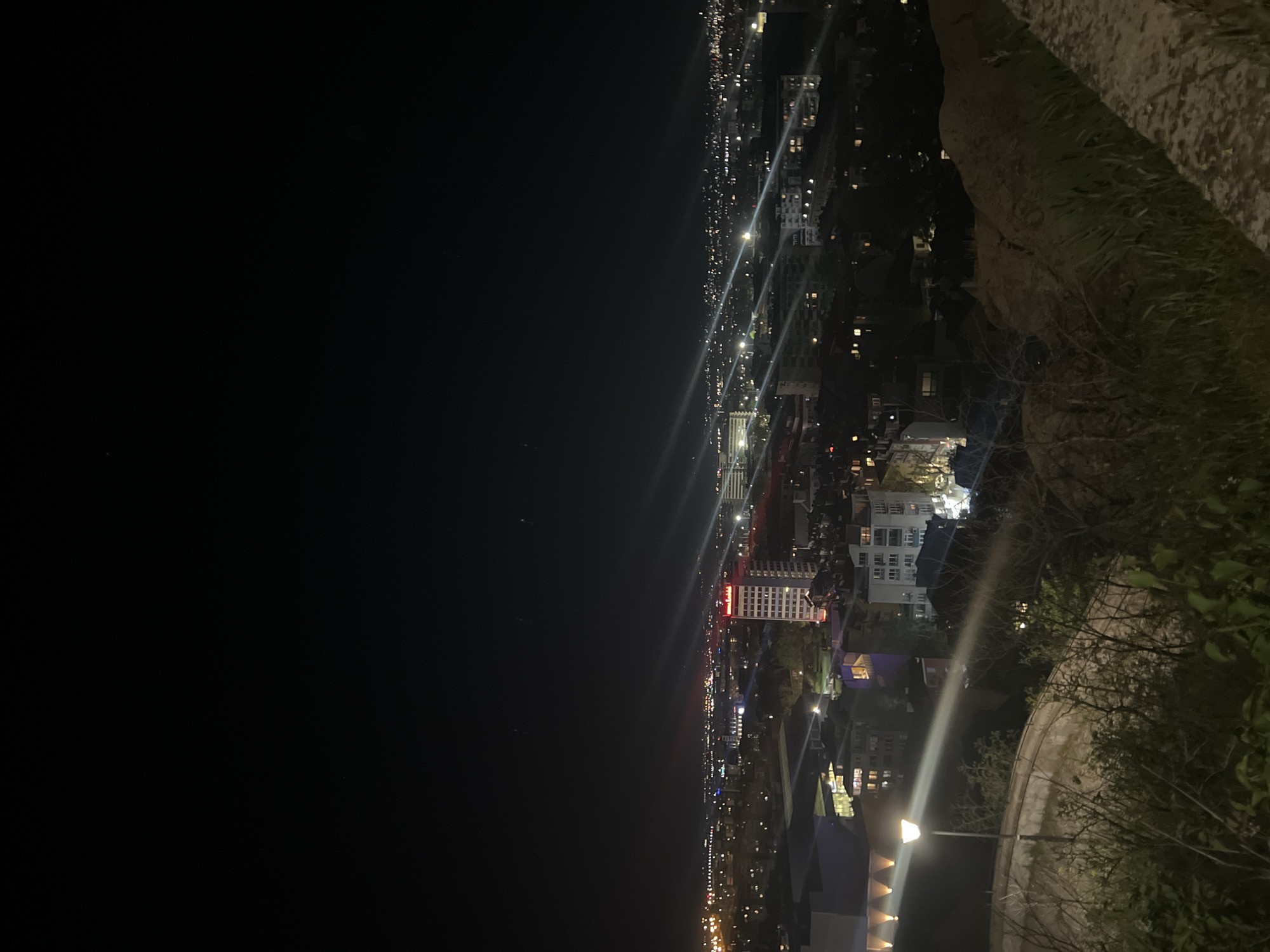 The image depicts a nighttime aerial view of Plovdiv, Bulgaria, taken from a high vantage point, possibly Nebet Tepe. The foreground shows vegetation and a stone wall. The city below is illuminated by streetlights and building lights. The horizon is defined by the skyline of the city, punctuated by buildings. The sky is dark.