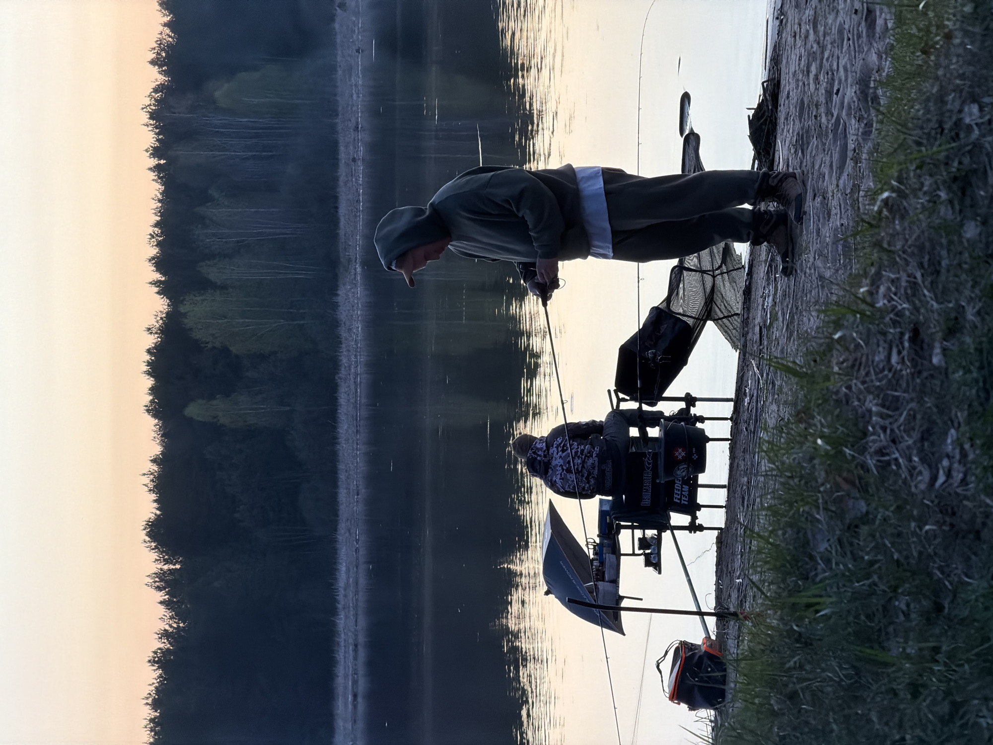Two people are fishing on a riverbank at dawn or dusk. One person is standing, holding a fishing rod and looking down at the water. The second person is sitting on a large fishing chair with numerous attachments. A fishing umbrella is set up beside them. Fishing tackle and bags are scattered nearby. The river reflects the trees and the sky. The location is likely near a body of water in Chaykovsky, Russia, based on the vegetation and the prevalence of fishing in Russian culture.