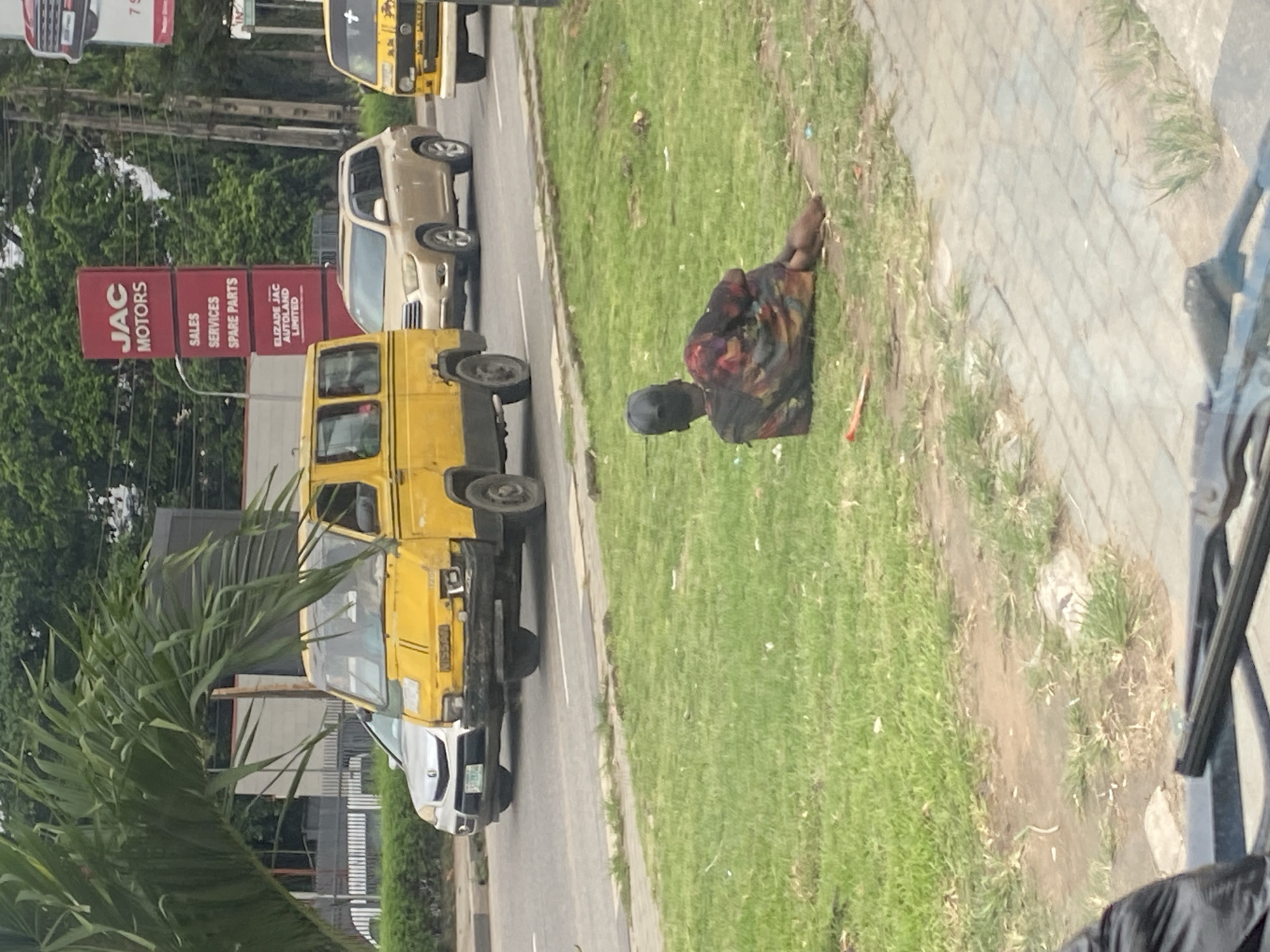 A person wearing a black baseball cap and a multi-colored shirt is lying on their side on a grassy area. The person is facing away from the camera, with their head resting on their arm. The grassy area appears to be a median strip on a road in Ikeja, Nigeria. Several vehicles, including yellow minibusses and a beige SUV, are driving on the road. A red sign with white text reading "JAC MOTORS" and listing services like "SALES", "SERVICES", and "SPARE PARTS" is visible in the background. Trees and buildings are also present in the background, suggesting an urban environment. The overall scene depicts a street view with a person resting on the roadside amidst traffic.