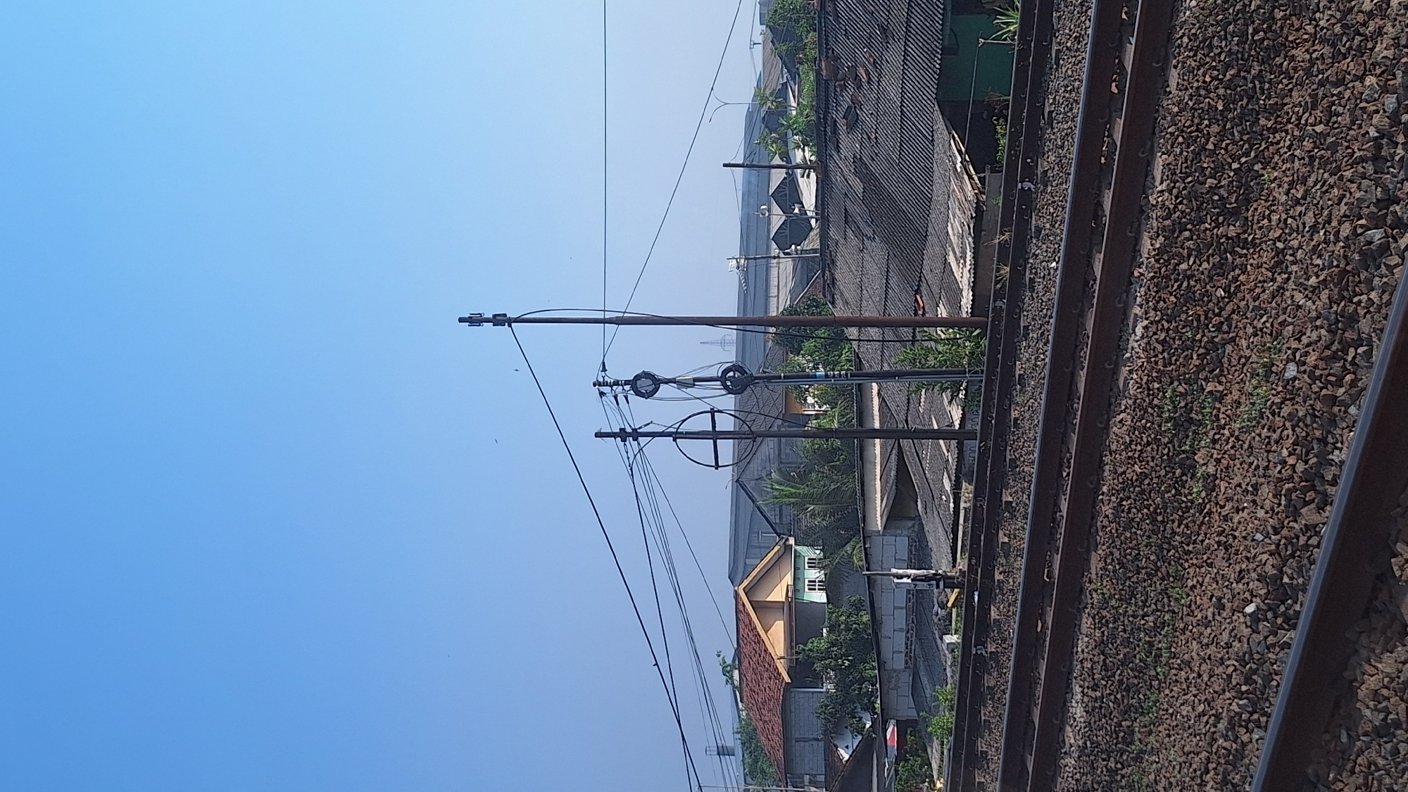 The image depicts a clear, sunny day in Waringinjaya, Indonesia, with a view over a residential area and railway tracks. In the foreground, multiple sets of parallel railway tracks are visible, laid on a bed of dark brown ballast. Beyond the tracks, a row of single-story houses with predominantly corrugated gray roofs stretches across the middle ground. Some of these houses feature brick walls, while others have lighter colored facades. Trees and vegetation are interspersed between the buildings.
In the background, a large, industrial-looking building with a gray roof and some structures with transparent or light-colored roofing materials are visible. A tall, slender electrical tower stands prominently on the right side of the image, supporting numerous electrical wires that crisscross the sky. Another pole with electrical equipment, including circular insulators, is positioned to the left of the electrical tower. The sky is a uniform, bright blue with a few faint specks that may be distant birds or small aircraft. The scene is devoid of any visible people. The overall impression is of an urban or semi-urban environment with infrastructure and residential housing under a clear sky.