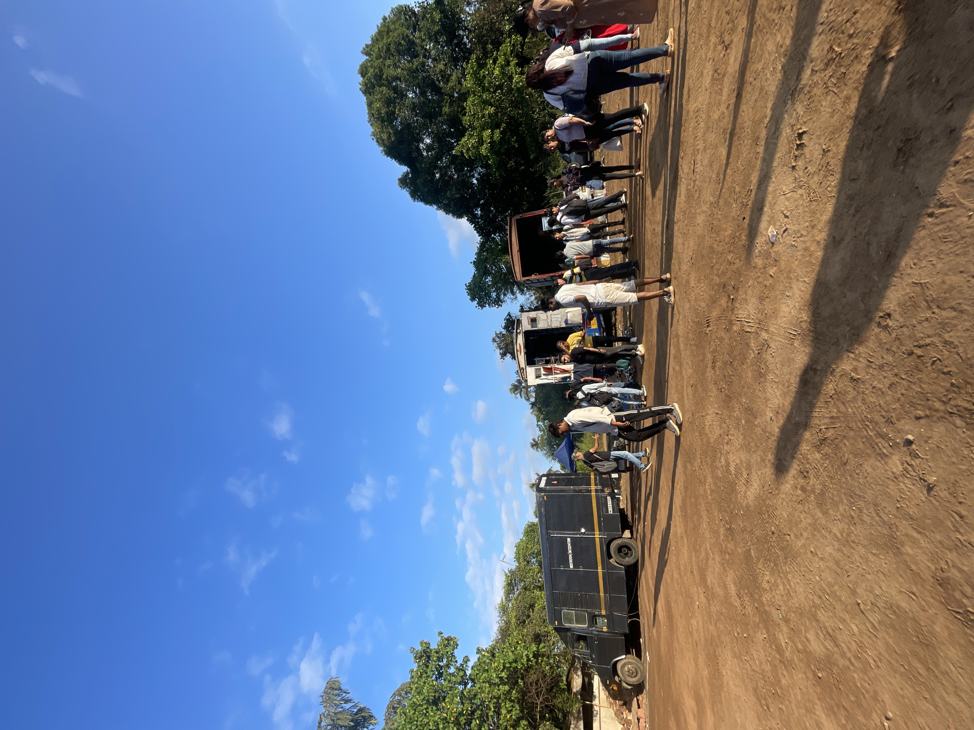 The image shows a group of people lined up near two vehicles on a dirt or sandy ground in Ashvem, India. The sky is blue with scattered clouds, and trees are visible around the scene. A black truck is parked on the left, and another vehicle (potentially a food truck or service vehicle) is positioned further up the line of people. The people appear to be standing or waiting. The overall impression is of an outdoor event or gathering.