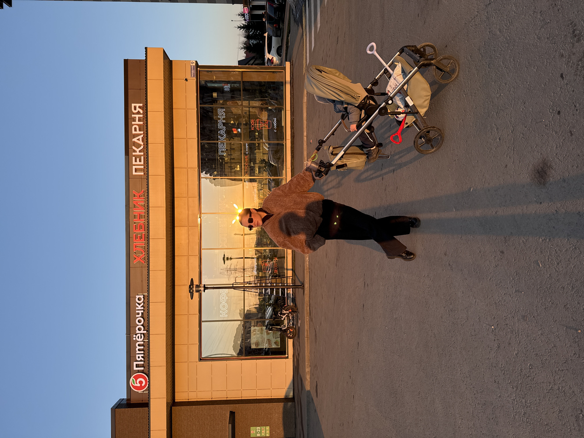 An adult female stands next to a beige stroller containing an infant on an asphalt surface in front of a commercial building. The location is Triumfalnaya Street, 9, Odintsovo, Russia, identified by the street sign reflected in the building's glass and the "Пятёрочка" (Pyaterochka) supermarket and "ХЛЕБНИК ПЕКАРНЯ" (Khlebnik Bakery) signage. The scene is illuminated by late afternoon or early evening sunlight, casting long shadows.
The female wears a brown faux-fur jacket, black pants, black boots, a black crossbody bag, and sunglasses. Her left hand rests on the stroller's handle. An infant, dressed in blue attire, occupies the stroller seat. A red toy shovel and a white object are visible in the stroller's lower storage compartment.
The single-story building features a light-brown facade with large reflective glass windows. Reflections show an electric scooter, a metal ladder, power lines, and internal signage including "КОФЕ" (Coffee) and "ПЕКАРНЯ" (Bakery). A dark multi-headed streetlight stands in front of the building. Parked vehicles and trees are visible in the background to the right. The sky is clear blue.