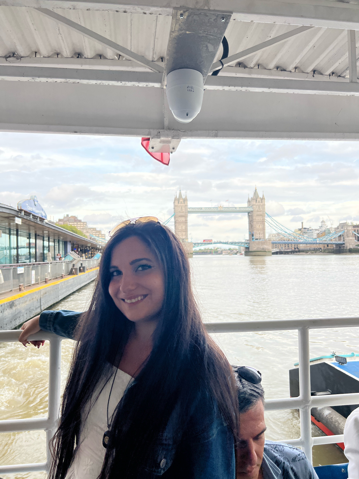 A woman with long, dark hair and dark eyes smiles at the camera, wearing a white top and a dark denim jacket. She is leaning on a white railing on a boat. Behind her, the River Thames flows under a cloudy sky. In the distance, Tower Bridge, a prominent landmark in London, United Kingdom, is visible. The riverbank features a station or terminal with a long building and glass windows. Another person's head and shoulder are visible to the right of the woman, partially obscured. A white ceiling structure with a white dome-shaped object is visible at the top of the frame.