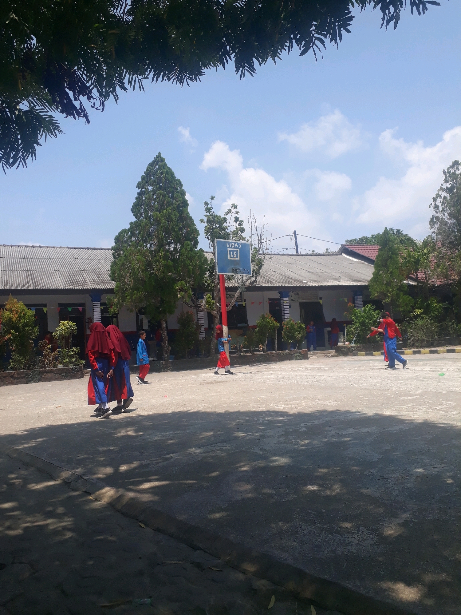 This image depicts an outdoor scene on a sunny day, likely within the grounds of a school in Pangkalpinang, Indonesia. In the foreground, a paved courtyard area is visible, with shadows cast by trees on the left side of the frame. A basketball hoop with a blue backboard and red pole is present in the mid-ground.
Several individuals are present. On the left, two young people wearing red headscarves and matching red and blue outfits are walking away from the camera. Further back and to the right of the basketball hoop, one person in a red shirt and blue pants is walking, and another individual in a blue shirt and red pants is also visible in the background, appearing to be engaged in an activity. Two children in blue shirts and red pants are standing near the school building.
The background features a long, single-story building with a grey, corrugated metal roof and white walls. There are trees and greenery surrounding the building and on the periphery of the courtyard. The sky is blue with scattered white clouds. The overall impression is of a schoolyard activity or break time.