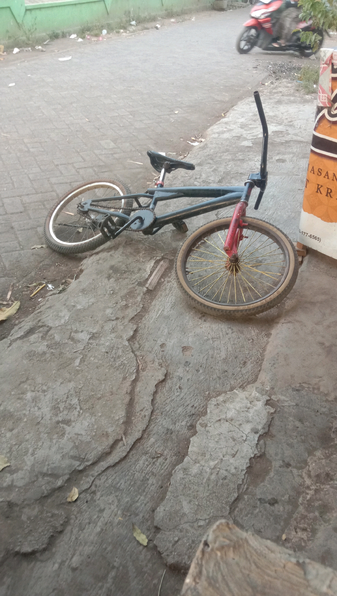 A black BMX bicycle is lying on its side on a cracked concrete surface in the city of Ngempit, Indonesia. The bicycle's front wheel is pointing upwards and to the right, while its back wheel is on the ground. The bicycle is dark, with a red front fork and a black frame. The seat is black, and the handlebars are black and curved. The bicycle is positioned on a concrete walkway adjacent to a paved road. In the background, a red motorcycle is parked, and there is a green wall and some scattered debris. A white and orange sign is partially visible to the right of the bicycle.