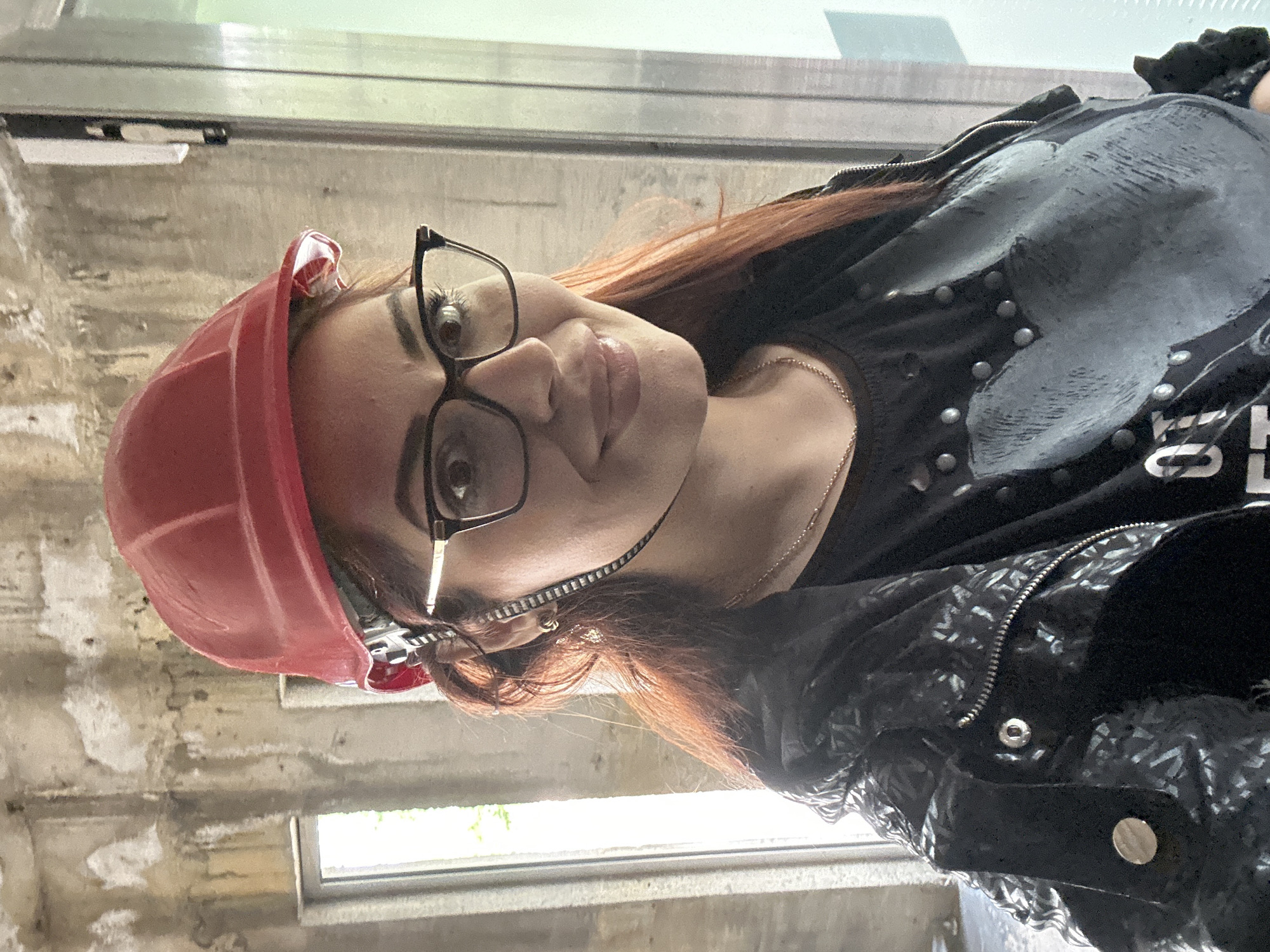 A young woman with auburn hair is wearing a red hard hat and black-rimmed eyeglasses. She is wearing a black top and a black leather jacket. She is standing near a concrete wall with what appears to be a window. The location is Gara Oreshets, Bulgaria.