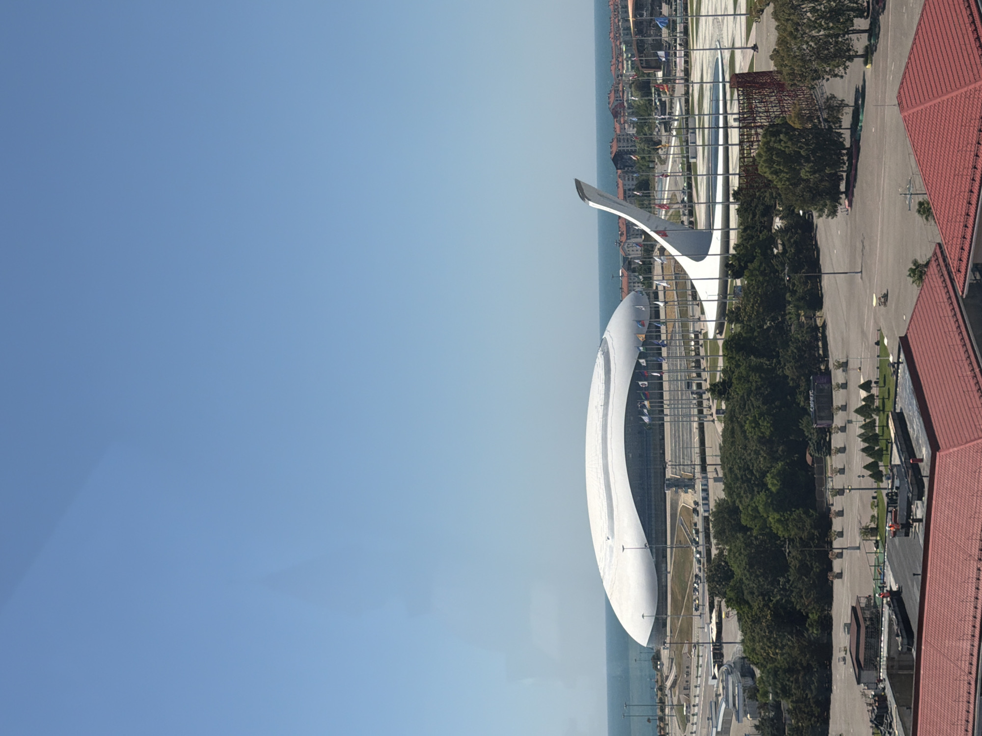 The image shows a wide view of a city area with a large, white, modern building with a curved roof in the background. In front of this building, there is a tall, white sculpture resembling a flame. Surrounding these structures are many flags on poles. The sea is visible in the distance behind the buildings and sculpture. The foreground shows several red-tiled roofs of buildings, some trees, and paved areas. The sky is clear and blue. The location is identified as Sirius, Russia. There are no clearly discernible people or specific actions taking place, but the scene depicts an outdoor urban environment with architectural landmarks.