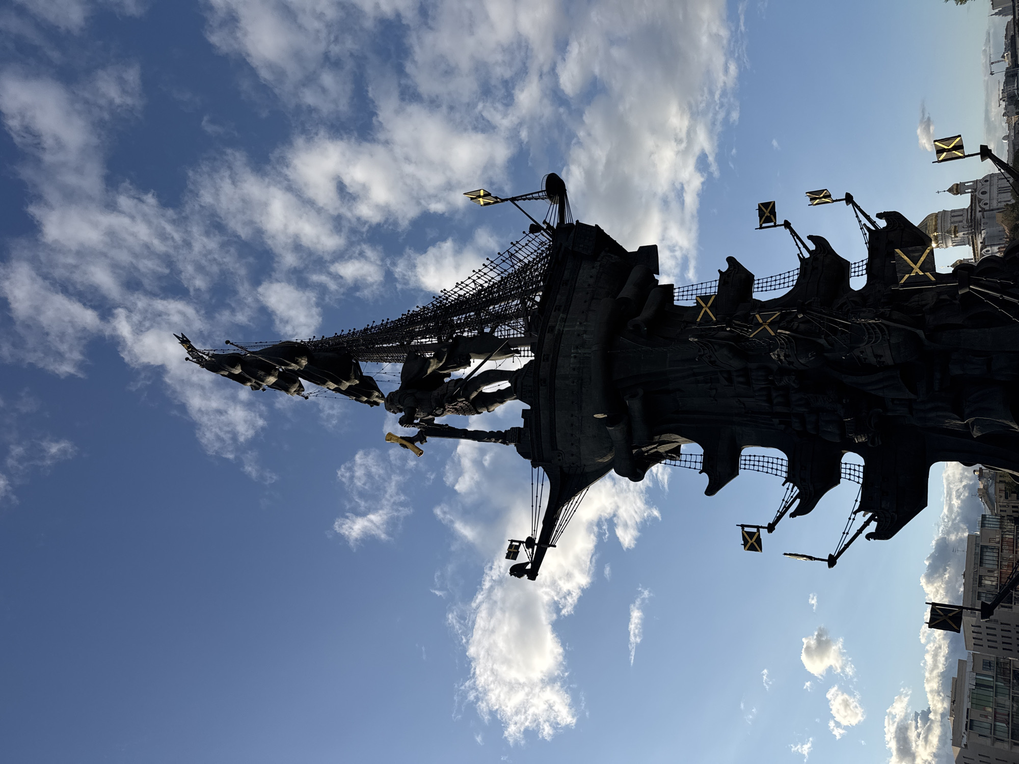 The image shows a low-angle, close-up, silhouette view of the Peter the Great Statue in Moscow, Russia. The statue, which depicts Peter the Great standing on a ship's hull with masts and sails, is silhouetted against a bright blue sky filled with scattered white clouds. The statue is a dark, monolithic structure, with intricate details of the ship's rigging, sails, and flags visible. Several yellow and black flags are attached to the ship's masts. In the background, the dome of a golden-domed building, likely a church, is visible. Modern buildings and construction cranes are also visible in the lower part of the image. The lighting suggests it is either early morning or late afternoon.