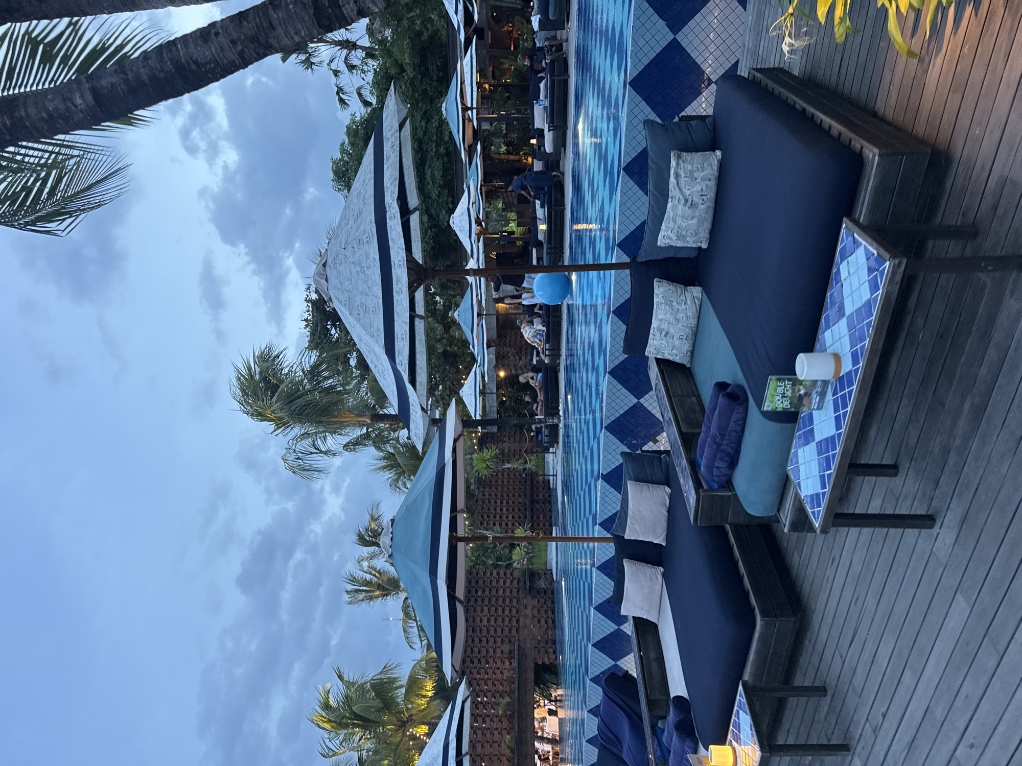 The image depicts a pool area in Kuta Selatan, Indonesia at dusk. The setting includes lounge chairs with blue cushions and blue and white patterned umbrellas lining the pool. The pool itself has a blue and white diamond pattern. A wooden deck surrounds the pool area. Palm trees are visible in the background and partially obscure the cloudy sky. People are present in the background, some possibly interacting in the pool area. A small table next to one of the lounge chairs holds a drink and a book.