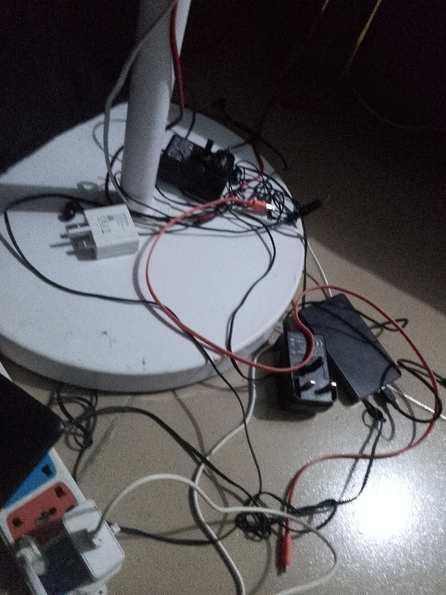 The image depicts a collection of electronic charging devices and cables scattered across a white, circular surface, possibly the base of a lamp, and the floor. The scene is dimly lit, with some glare visible on the floor's textured surface.
Specifically, there are several white and black power adapters plugged into what appears to be a power strip or directly into the floor outlets. Multiple charging cables, in black and red, are entangled around these devices. A black rectangular object, potentially a power bank or a smartphone, is also visible among the clutter. The overall impression is one of disarray, with cables haphazardly arranged.
The location of Jauro Yinu, Nigeria, is mentioned, but there is no visual information within the image that confirms this specific geographical context. The environment appears to be indoors, given the presence of electrical appliances and the floor's texture, but no specific architectural features or outdoor elements are discernible. There are no people or any interactions between subjects visible in the image.
