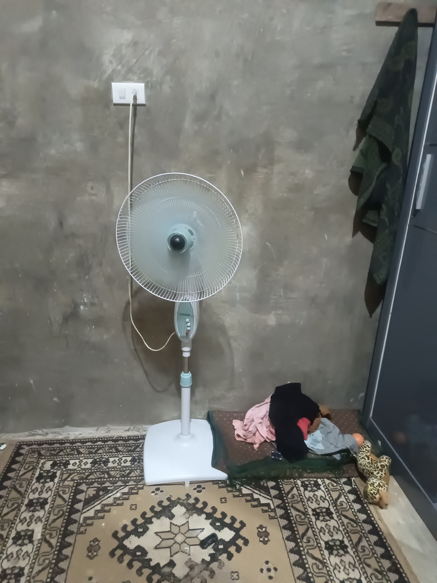 In a dimly lit room with concrete walls, a white pedestal fan stands to the left, plugged into a wall socket. A colorful patterned rug covers the floor in front of the fan. To the right, a pile of clothing, including a pink shirt and a black garment, is scattered on a brown mat. A small leopard-print stuffed animal is also visible near the clothing. A dark green towel or fabric hangs on the wall to the far right, next to a dark gray doorframe.
