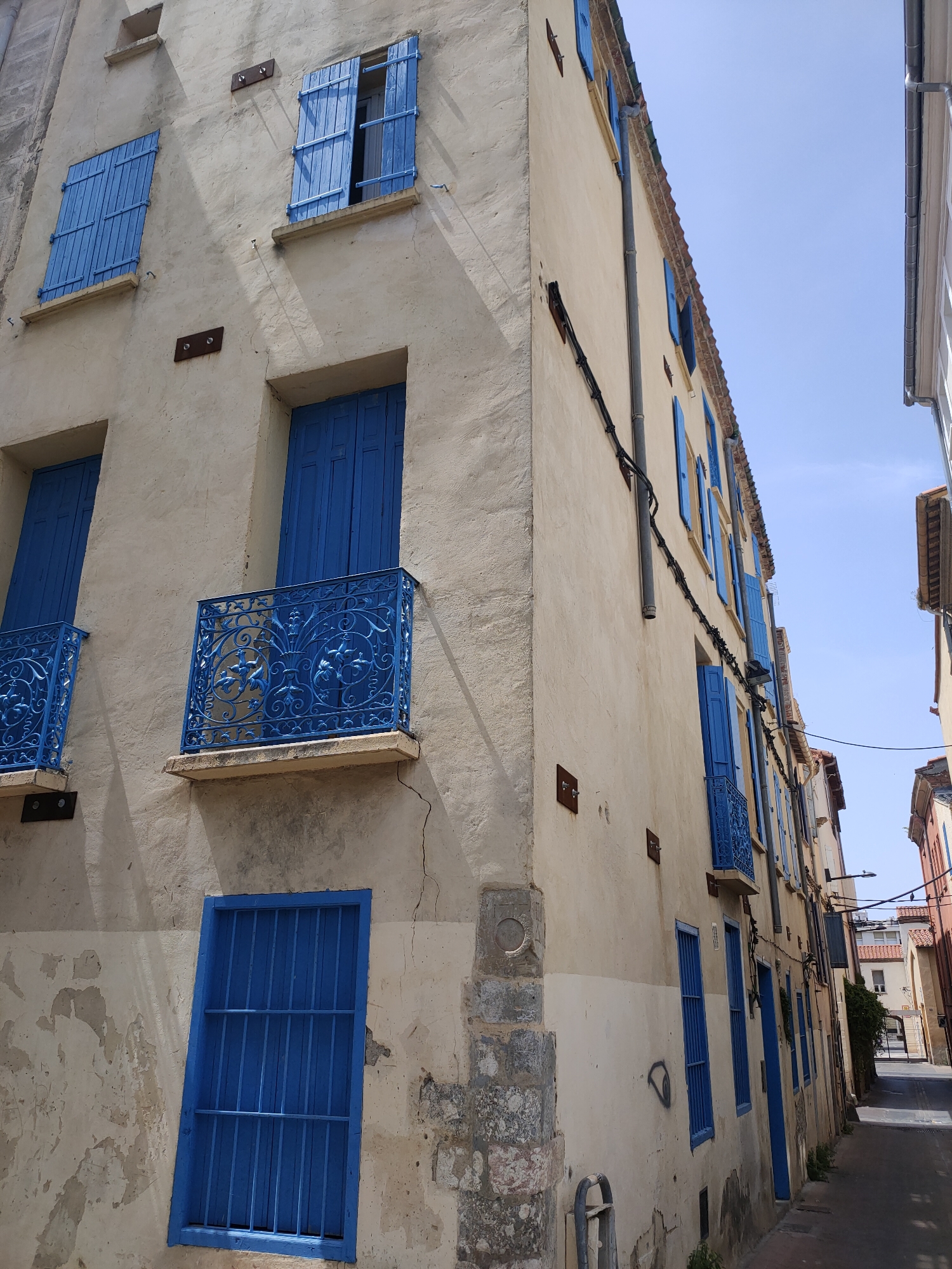 This is a low-angle, full shot photograph of a narrow street in Perpignan, France, likely in the historic district. The primary focus is the weathered, cream-colored facade of a multi-story building on the left, with a pronounced crack running vertically near the center. The building features numerous windows, most of which are covered by vibrant blue shutters. Some shutters are open, revealing dark interiors, while others are closed. Two small balconies with intricate blue wrought-iron railings are visible, one with a detailed floral motif. Below these balconies, two barred windows with similar blue frames are prominent. The building's exterior shows signs of age and wear, with peeling paint and exposed stonework.
The street itself is paved, and the building on the right side of the frame is partially visible, also with blue shutters. The sky above is clear and bright blue. Electrical conduits and pipes run along the side of the main building. The overall impression is of a sun-drenched, somewhat dilapidated but charming urban environment.