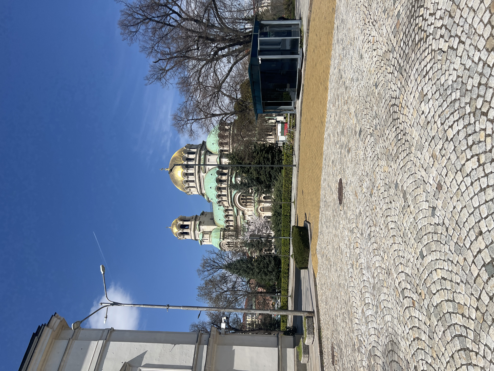 The image shows the Alexander Nevsky Cathedral in Sofia, Bulgaria. The cathedral's golden domes and green roof are visible against a clear blue sky. In the foreground, there is a cobblestone path, with a small structure on the right. A street lamp and part of a white building are visible on the left. Trees without leaves surround the Cathedral. A contrail is visible in the sky.