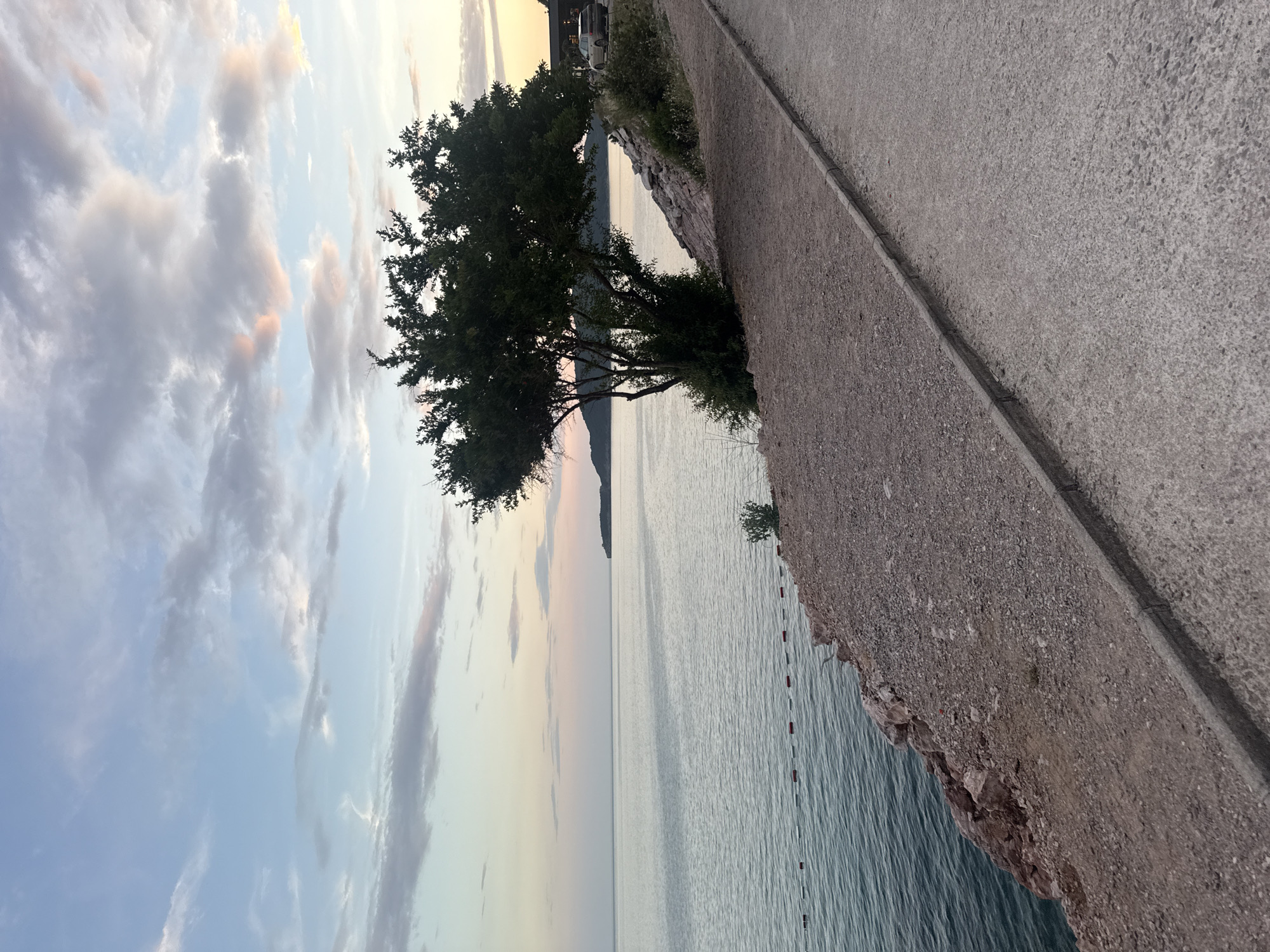 This image depicts a tranquil scene along the coast of Kamenovo, Montenegro, likely captured during either sunrise or sunset, given the soft, diffused light and pastel hues in the sky. The foreground is dominated by a textured, grey concrete pathway or promenade that runs diagonally across the frame, suggesting a developed coastal area. Adjacent to the pathway, a rocky embankment slopes down towards the sea.
The sea itself is calm, with gentle ripples reflecting the muted colors of the sky. A series of red and white buoys mark an area in the water, possibly a designated swimming zone or a boundary. On the left side of the image, a lone tree with dense foliage stands near the water's edge, its silhouette contrasting with the bright sky. In the distance, a faint outline of hills or mountains can be seen on the horizon, adding depth to the landscape.
The sky is a canvas of soft colors, with streaks of pale pink, orange, and yellow blending into a pale blue. Wispy clouds are scattered across the sky, catching the light and adding texture to the atmospheric perspective. There are no people or animals clearly visible in the scene. The overall mood is one of peacefulness and natural beauty, inviting contemplation. The lighting and the calm sea suggest a serene moment, perhaps the end of a day or the beginning of a new one, offering a sense of quietude.