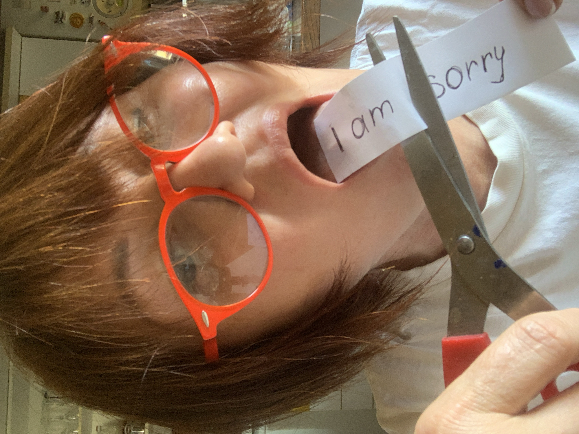 A close-up, overhead shot shows a woman's face, wearing bright orange, round-rimmed glasses and a white t-shirt. She has reddish-brown hair and her mouth is open, with a strip of paper in it that reads "I am sorry." She is holding a pair of scissors, with the blades poised to cut the paper. The background is blurred, but appears to be an indoor setting with shelves and bottles visible. The image does not provide any information about the location in San Quirico d'Orcia, Italy.